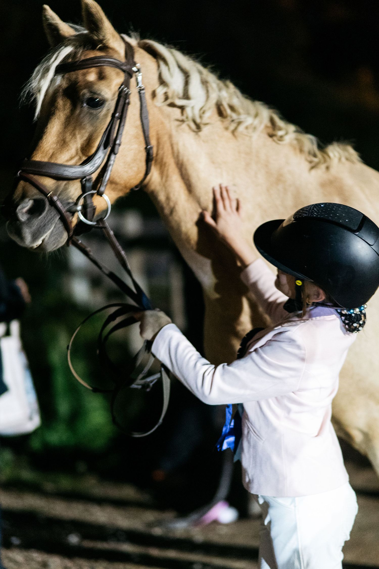 Young girl in black riding helmet gently petting a palomino horse wearing a bridle at an equestrian event.