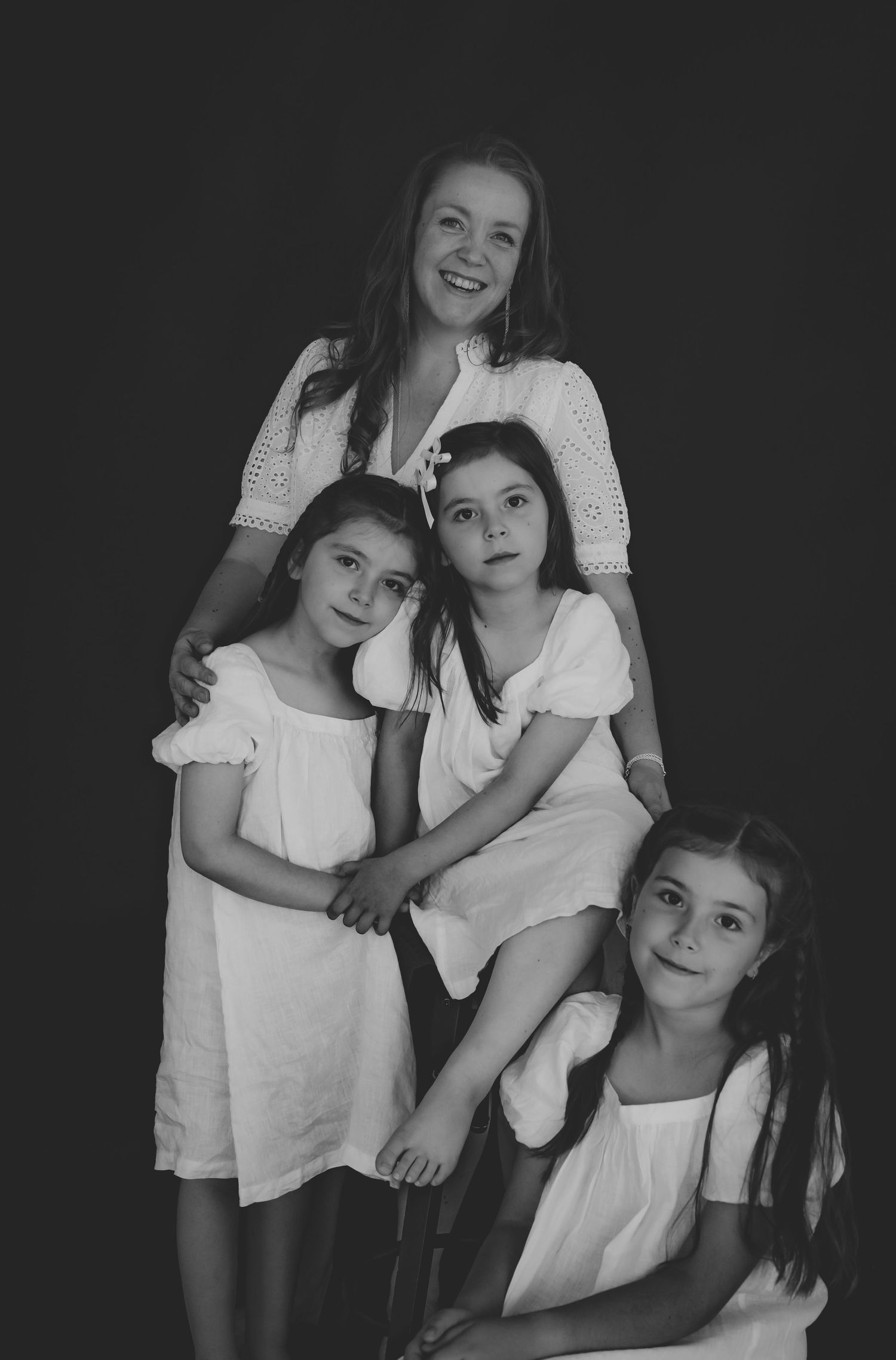 Black and white portrait of a smiling woman with three young girls in white dresses against a dark background.