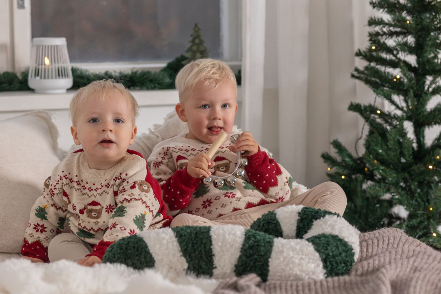 Two toddlers in matching Christmas sweaters sit on a cozy blanket beside a decorated tree, holding cookies near a striped candy cane pillow.