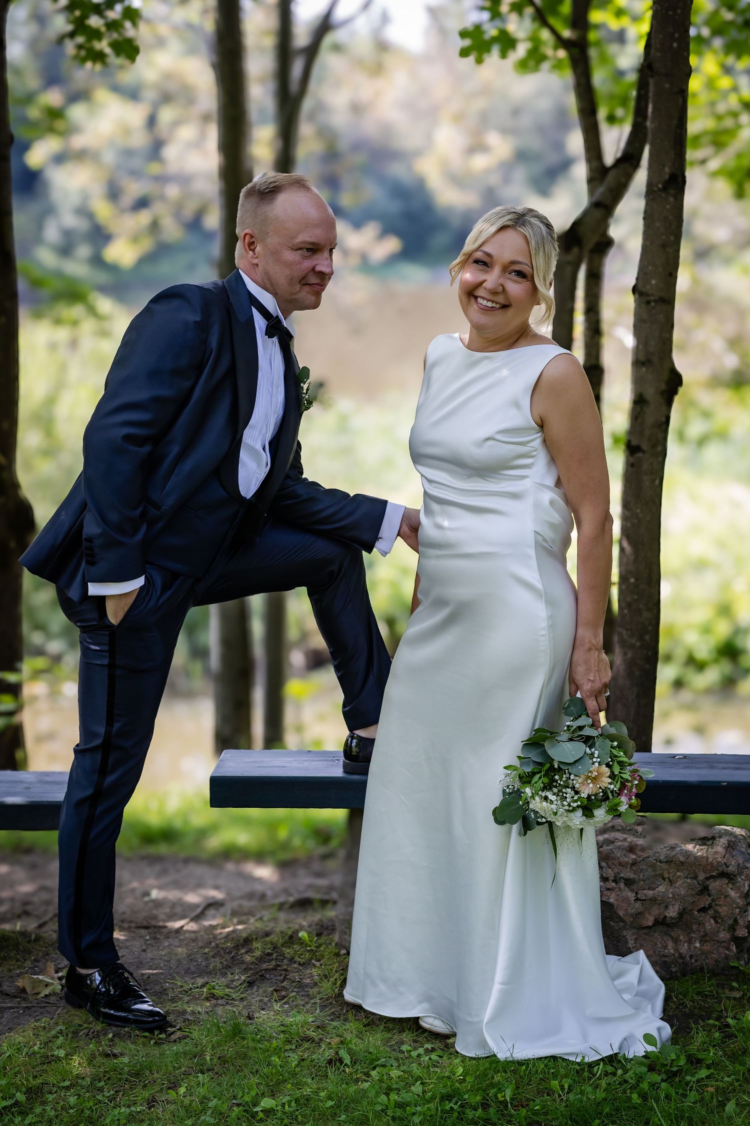 Bride in sleek white gown and groom in navy tuxedo posing playfully on bench in park setting.