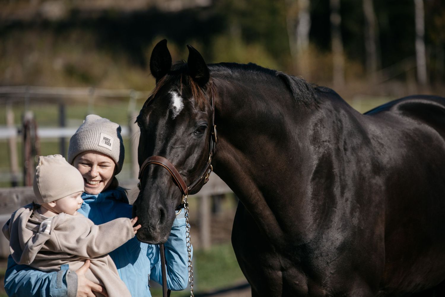 A smiling woman holds a baby while gently petting a dark brown horse outdoors at a sunny equestrian farm.