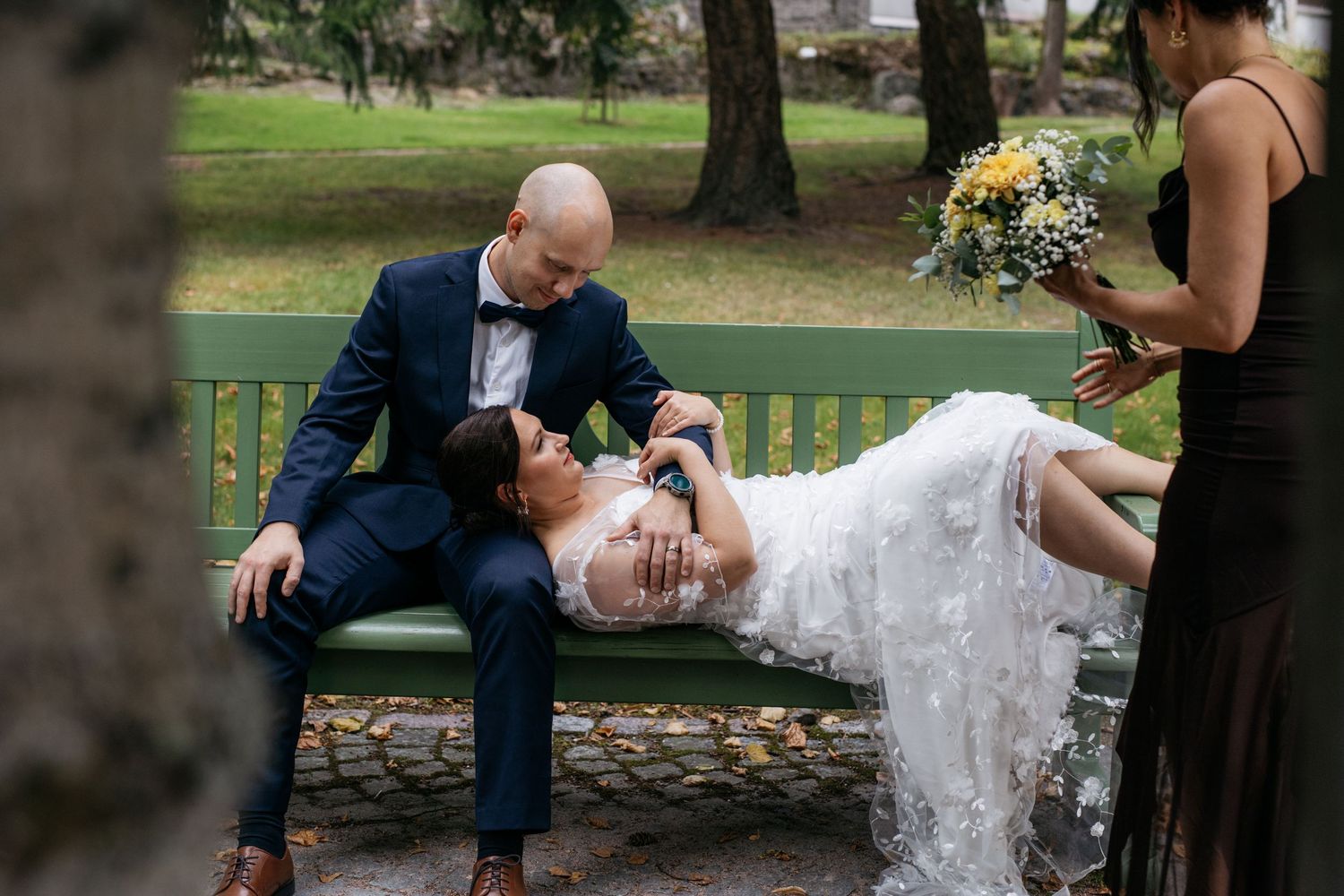 Bride lying across groom's lap on a green park bench while bridesmaid holds yellow bouquet nearby.