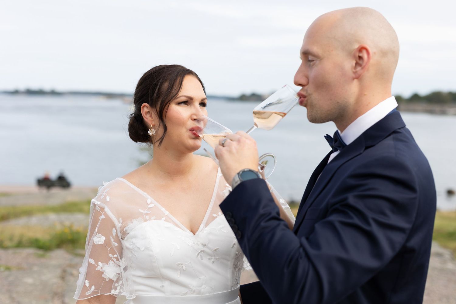 Bride and groom sharing champagne toast by waterside, bride in lace dress, groom in navy suit with bow tie.