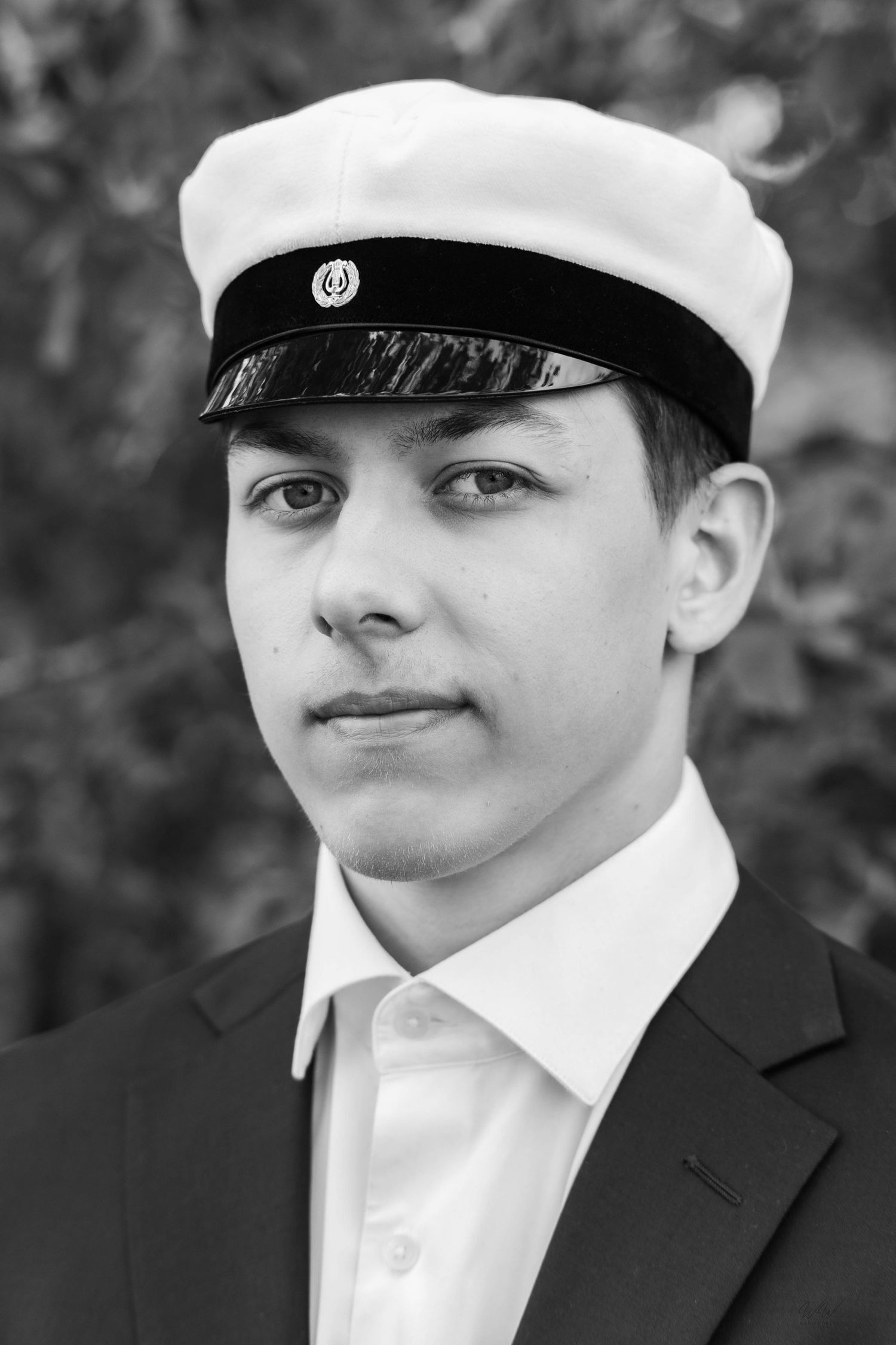 Black and white portrait of a young man in a white student cap with a black band, dark blazer and white collared shirt, outdoors.