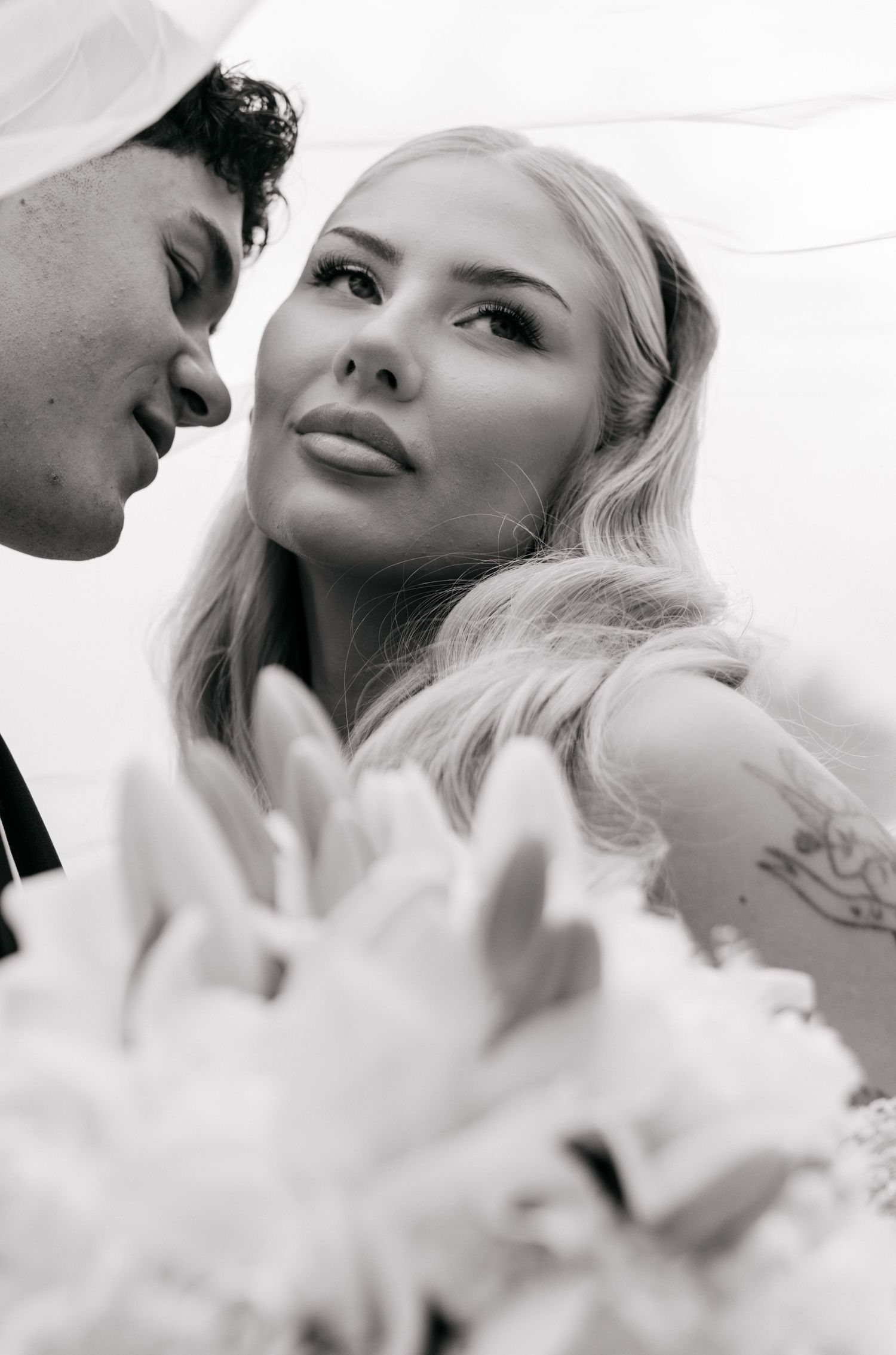 Black and white close-up of blonde bride gazing at groom under veil with blurred floral bouquet in foreground.