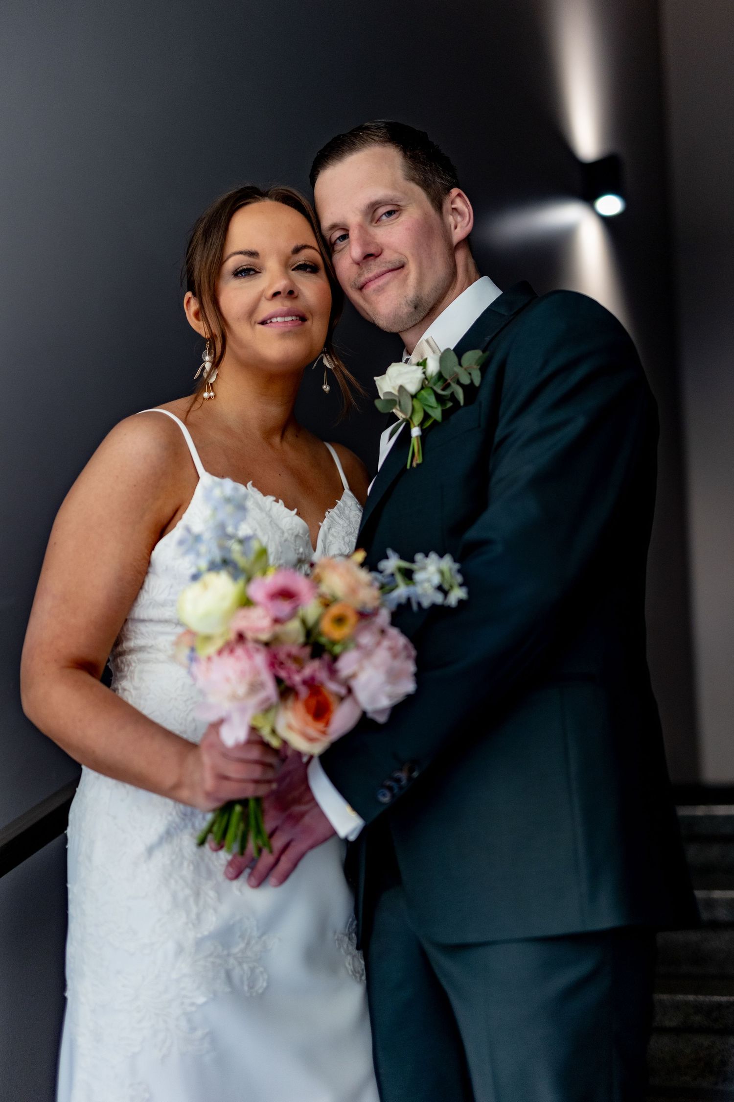 Bride in lace gown holding colorful bouquet poses with groom in dark suit on elegant staircase.