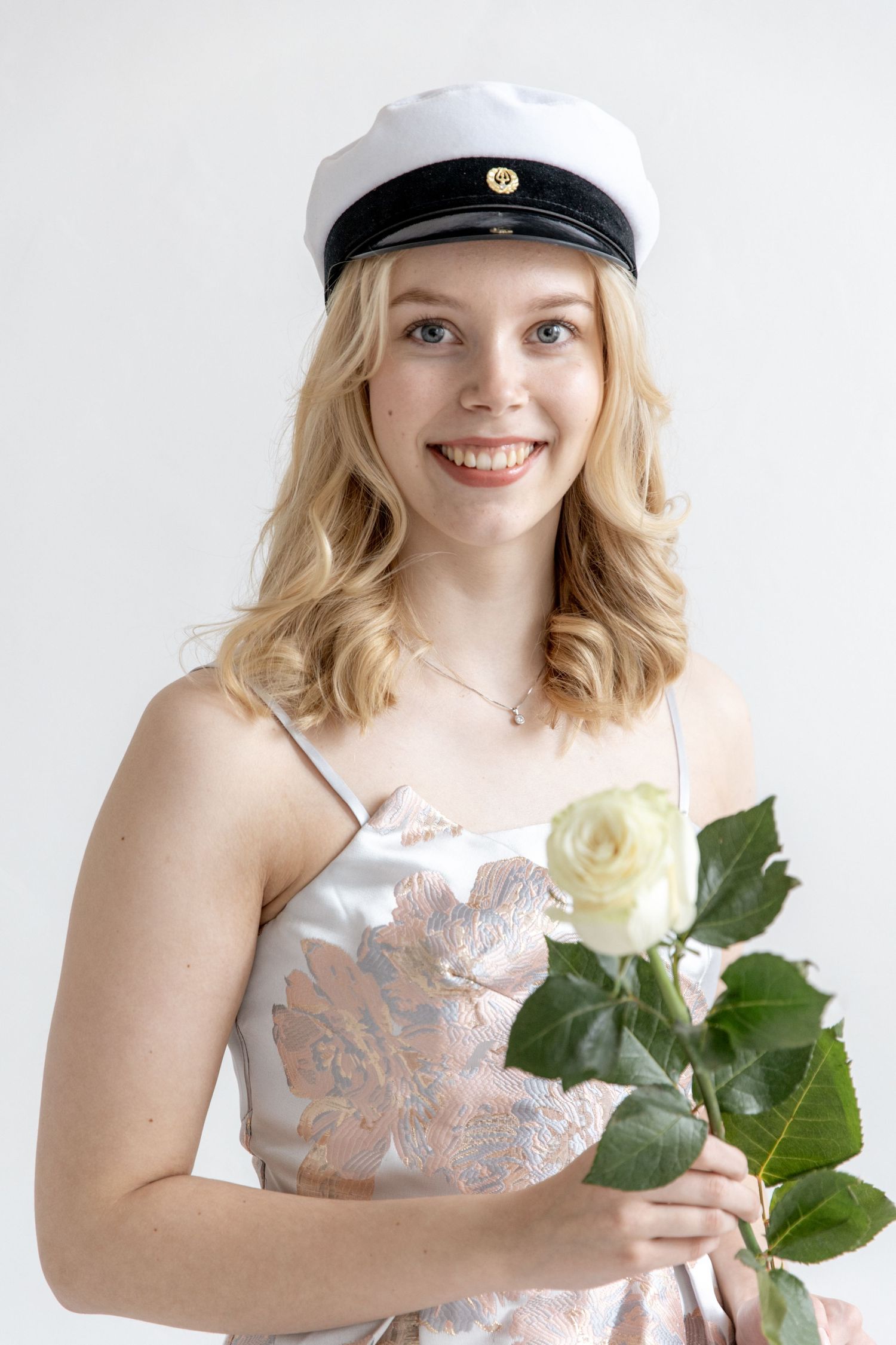 Smiling young woman in a white graduation cap and floral satin dress holding a white rose against a bright background.
