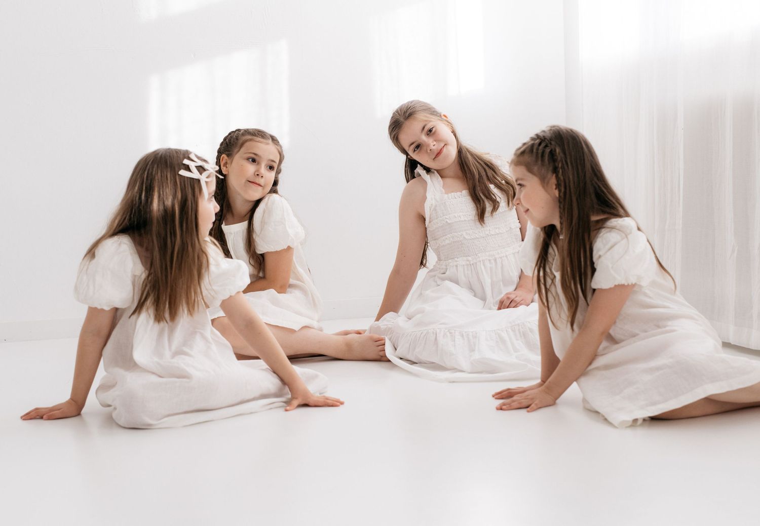 Four girls in white dresses sitting together on a white floor, smiling and interacting in a bright, airy setting.