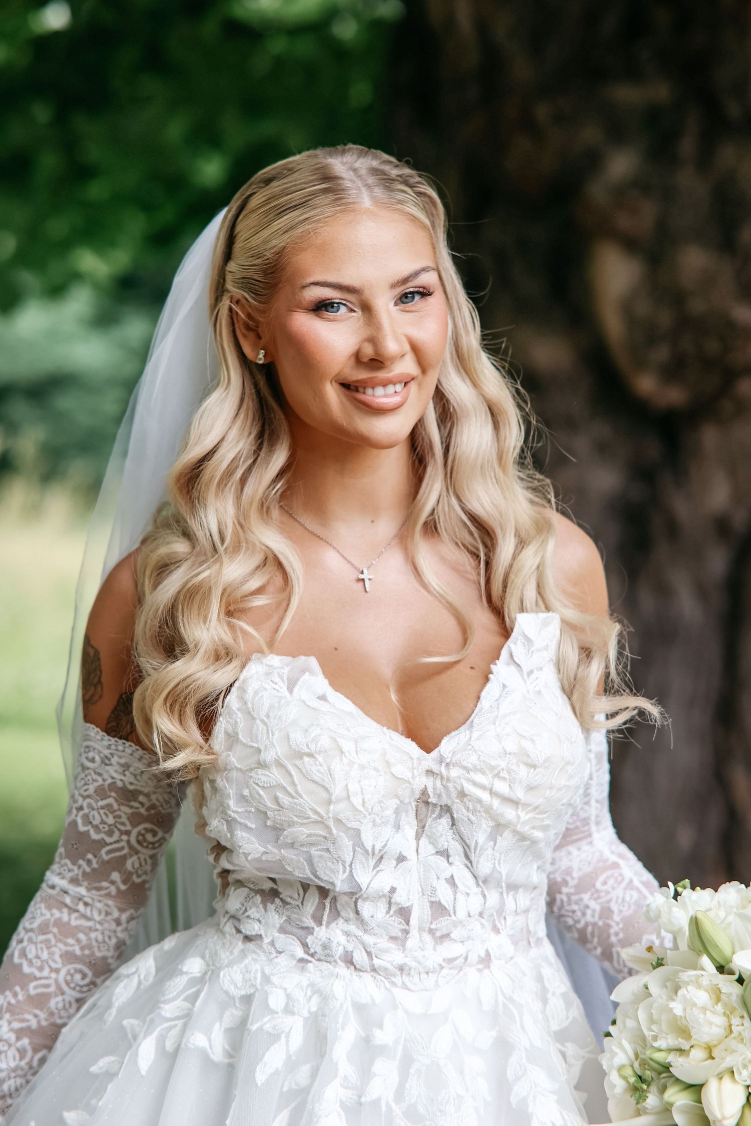 Smiling bride in white lace off-shoulder gown with veil and cross necklace holding bouquet outdoors.
