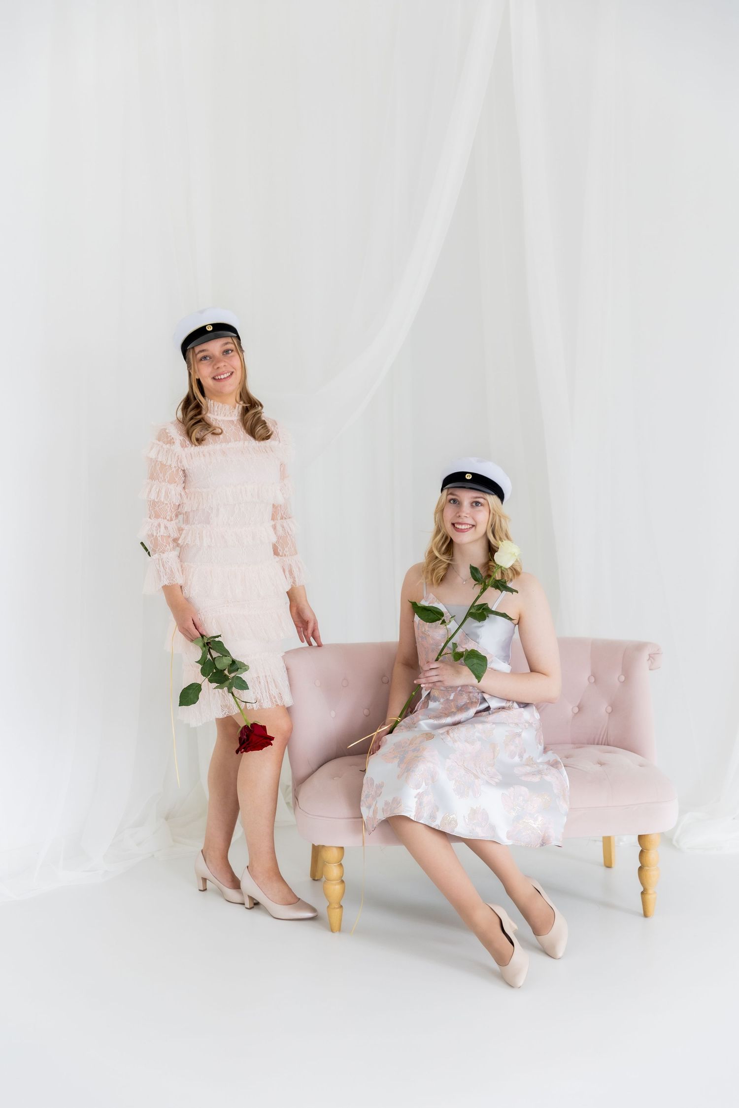 Two women in pastel dresses and white graduation caps hold red roses beside a pink tufted sofa against a soft white drapery backdrop.
