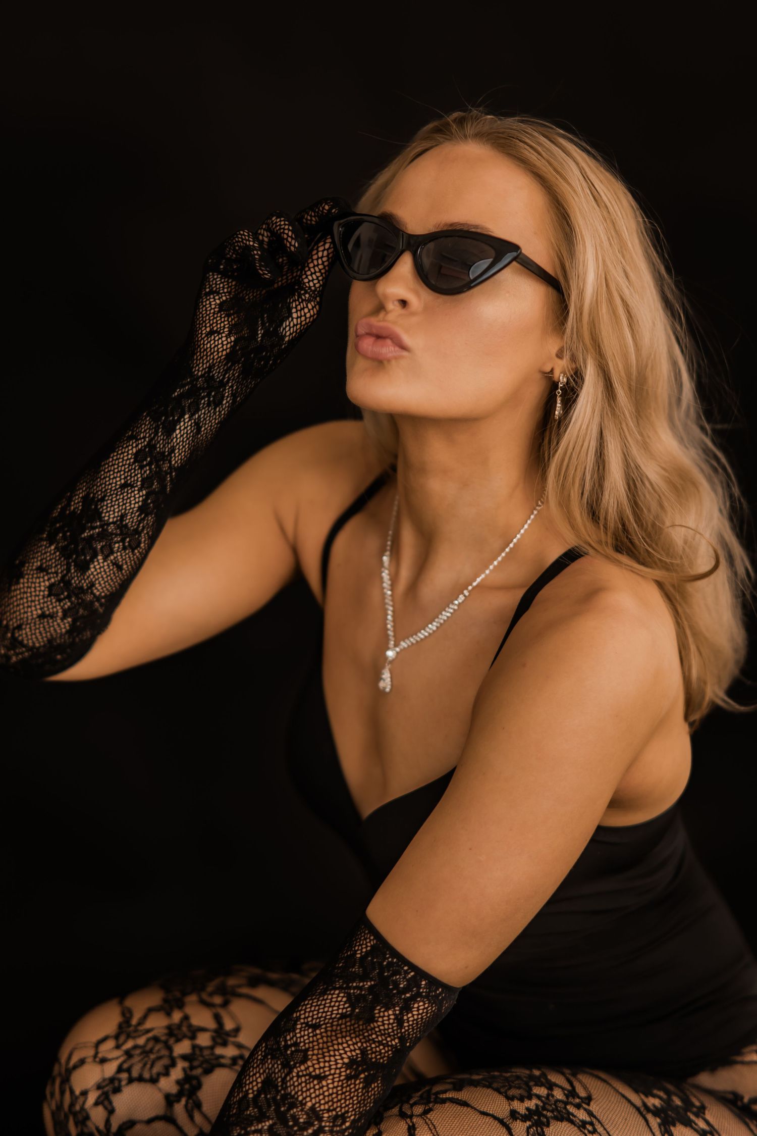 Blonde woman in black bodysuit with lace gloves, cat-eye sunglasses, and diamond necklace posing against dark background.