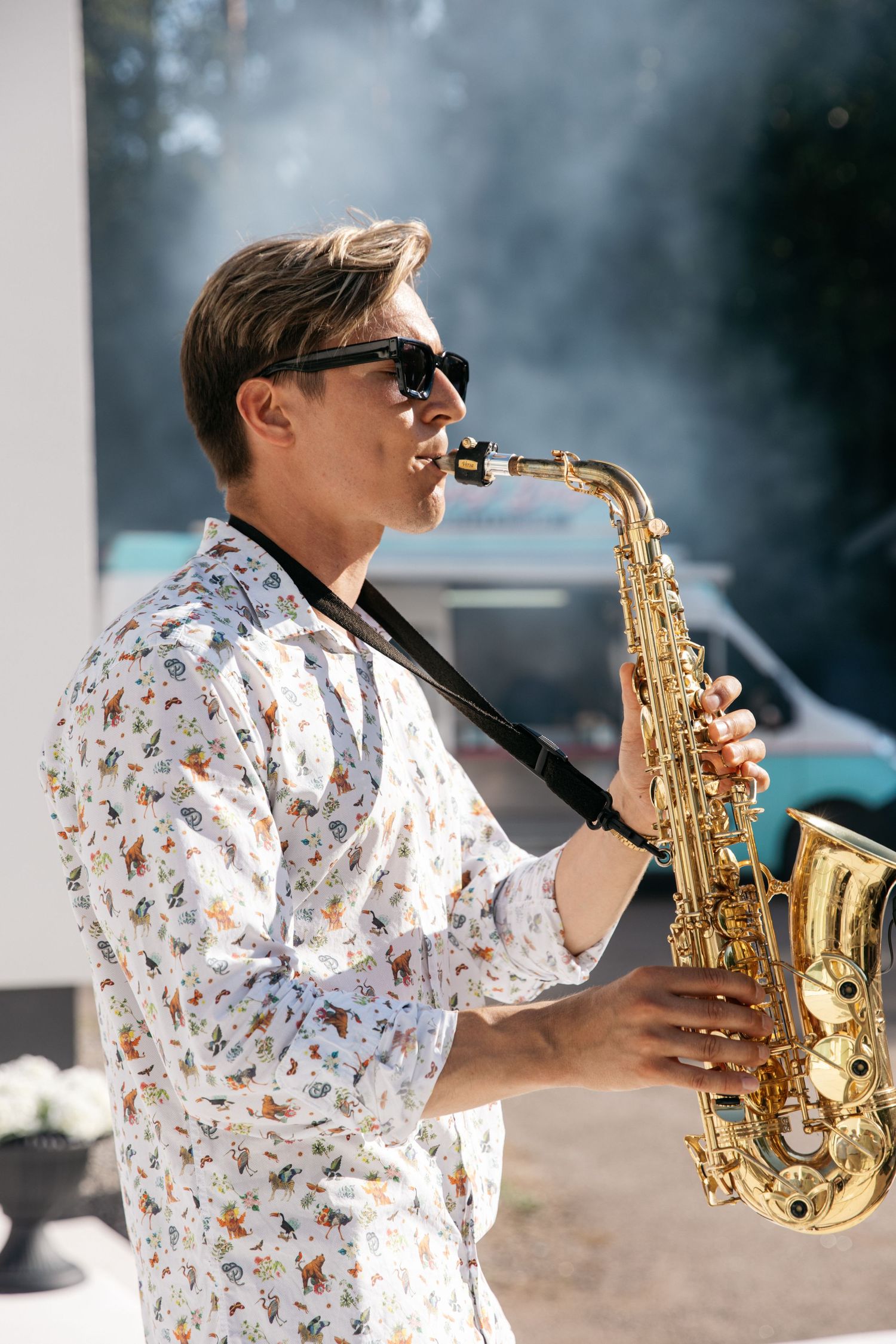 A musician in sunglasses and a patterned shirt plays a gold saxophone outdoors with smoke in the background.