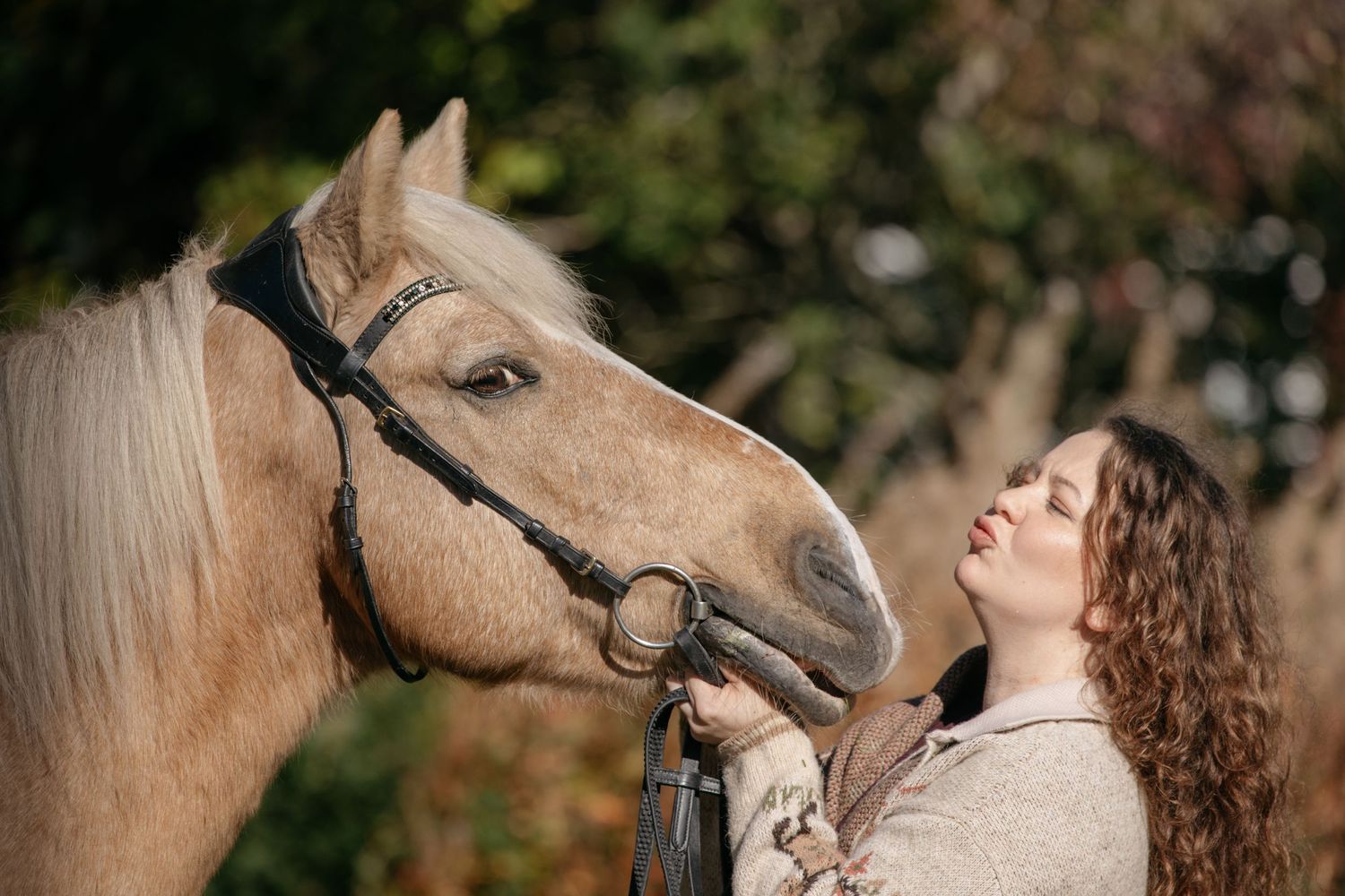 Young woman with curly hair gently holds the bridle of a palomino horse outdoors in soft natural light.