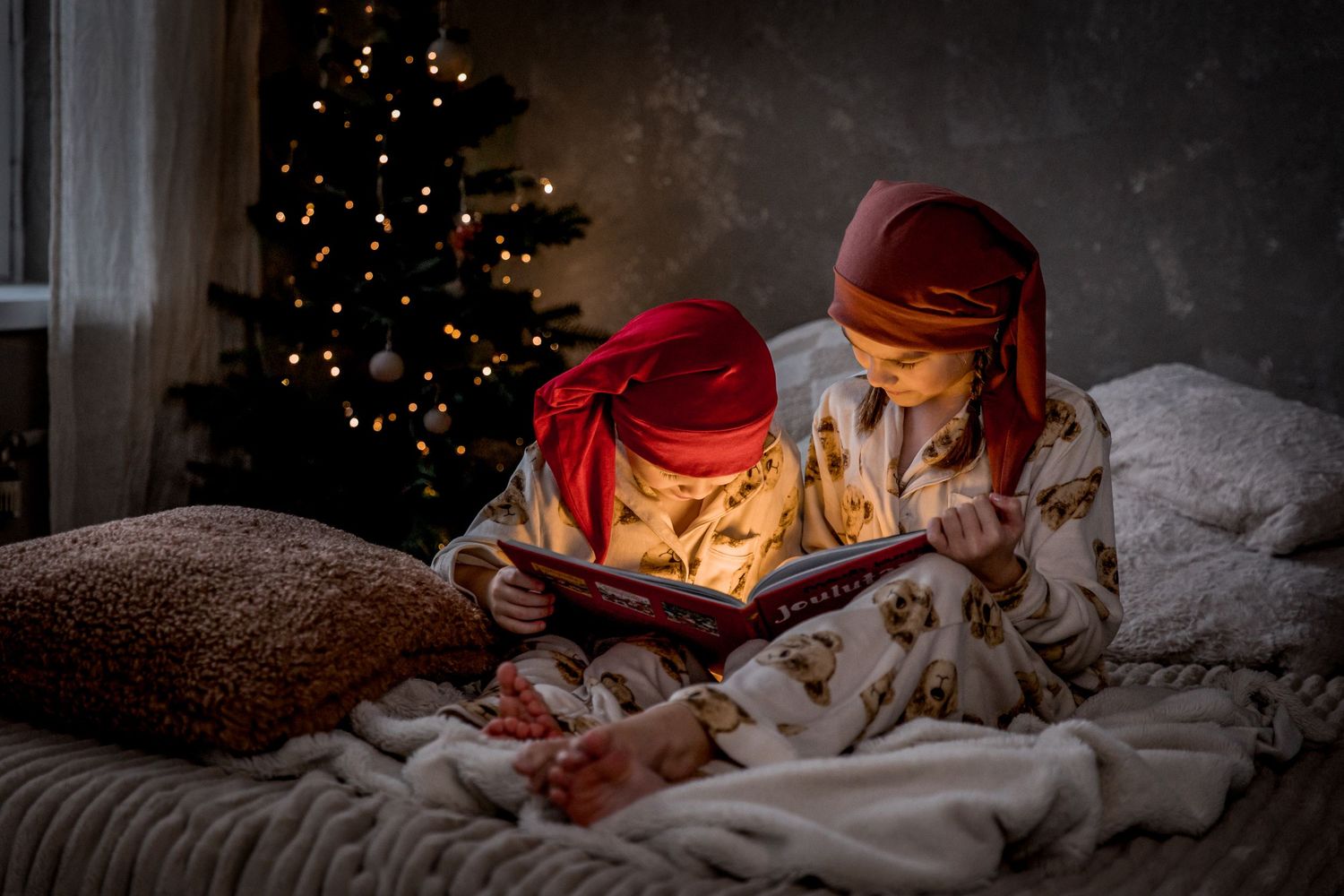 Two children in red head scarves and cozy pajamas read a glowing book together on a bed beside a lit Christmas tree.