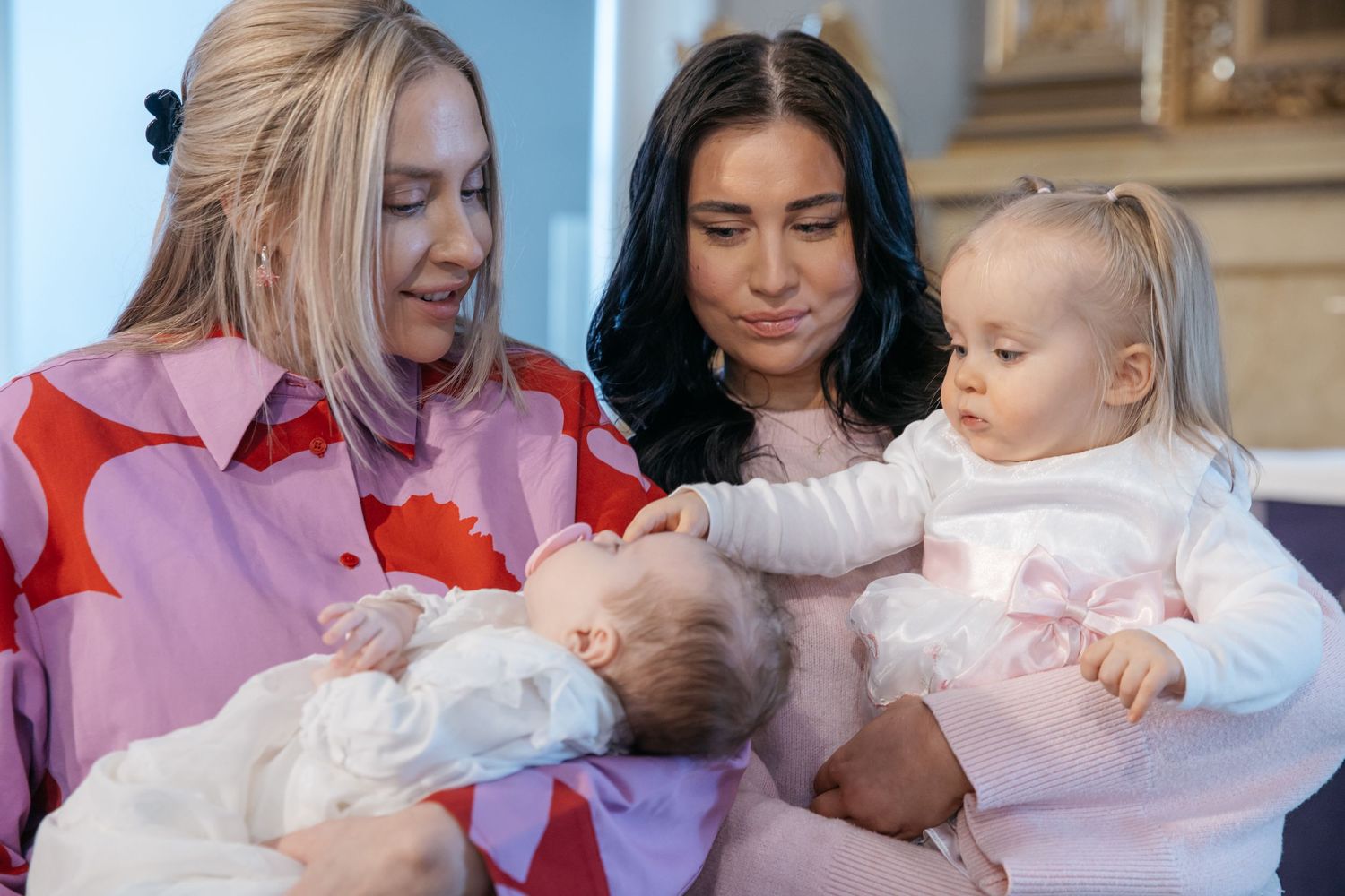 Two young mothers sitting together holding their babies at an indoor christening or family event.