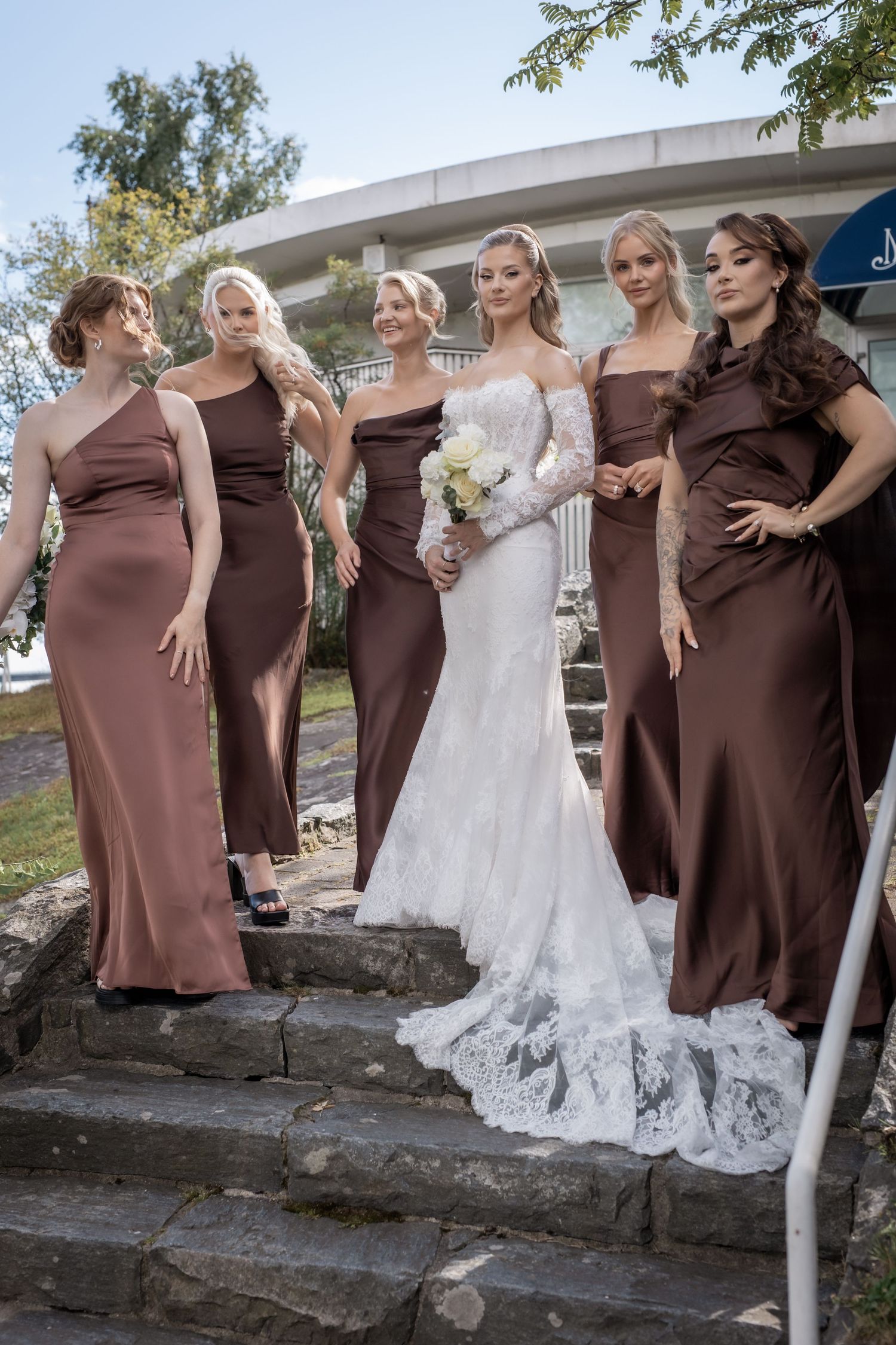 Bride in lace off-shoulder gown poses with five bridesmaids in brown satin dresses on steps.