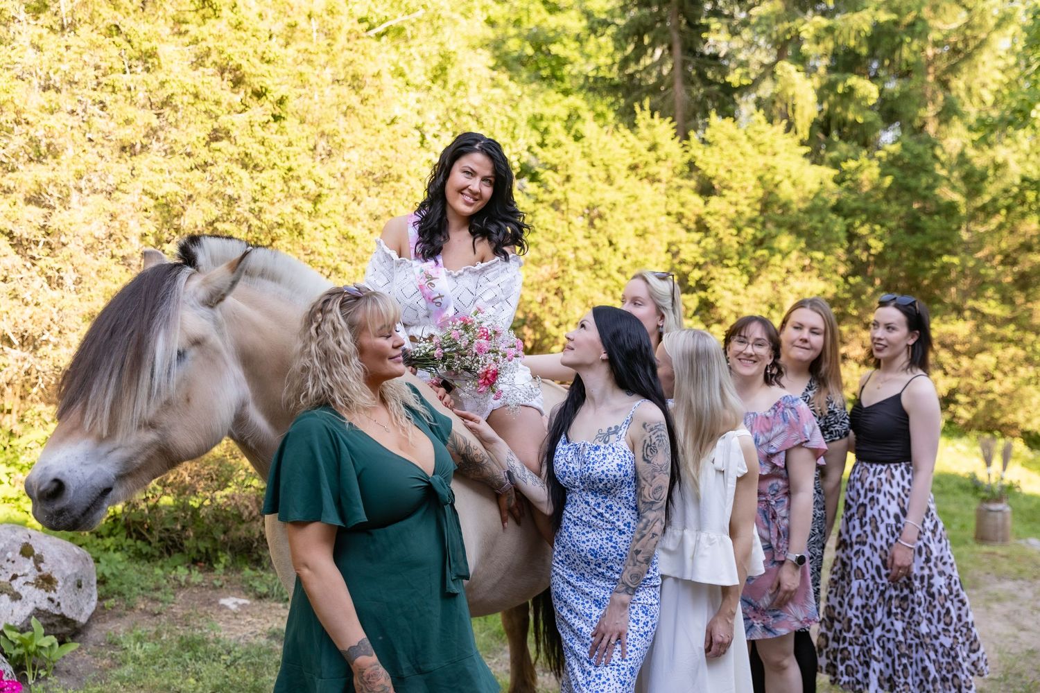 A smiling bride on horseback holds a bouquet surrounded by guests in a sunny outdoor forest setting.