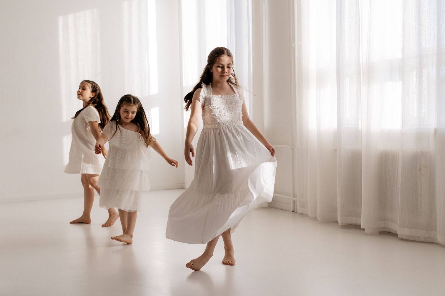 Three young girls in white dresses dance barefoot in a bright white room with sheer curtains.