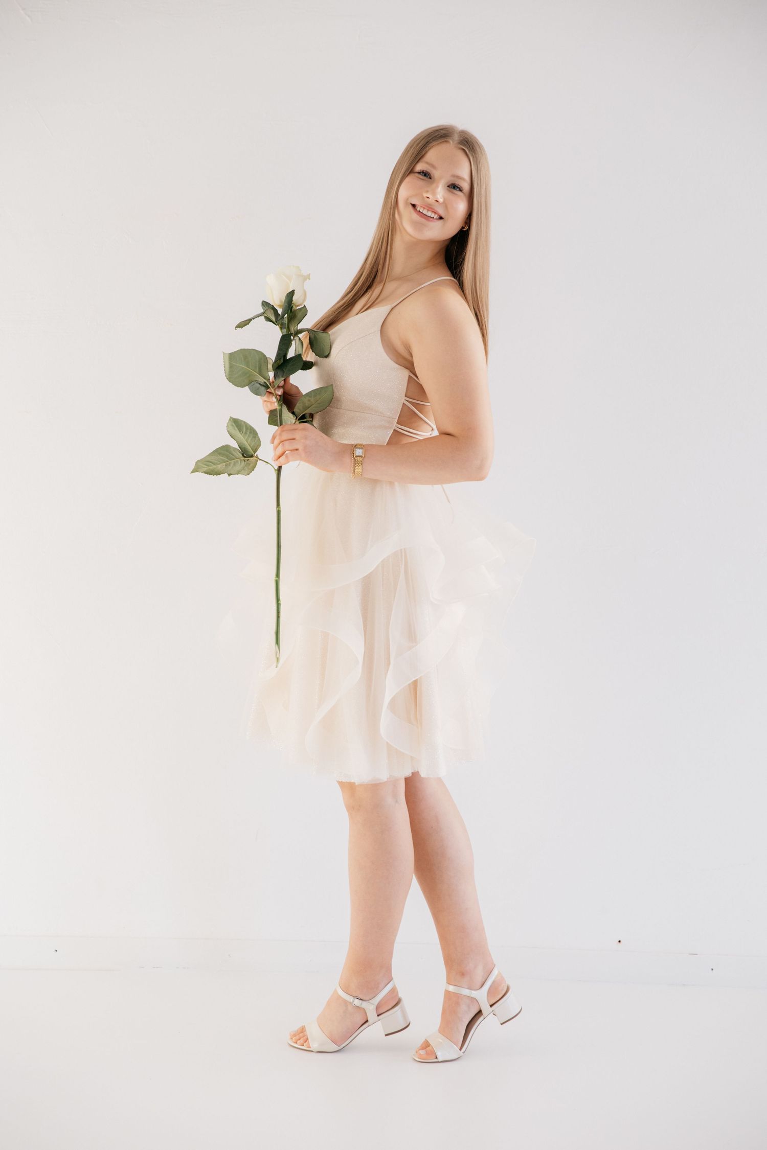 Smiling woman with long blonde hair in a cream tulle ruffled dress and white heels holding a white rose against a bright backdrop.