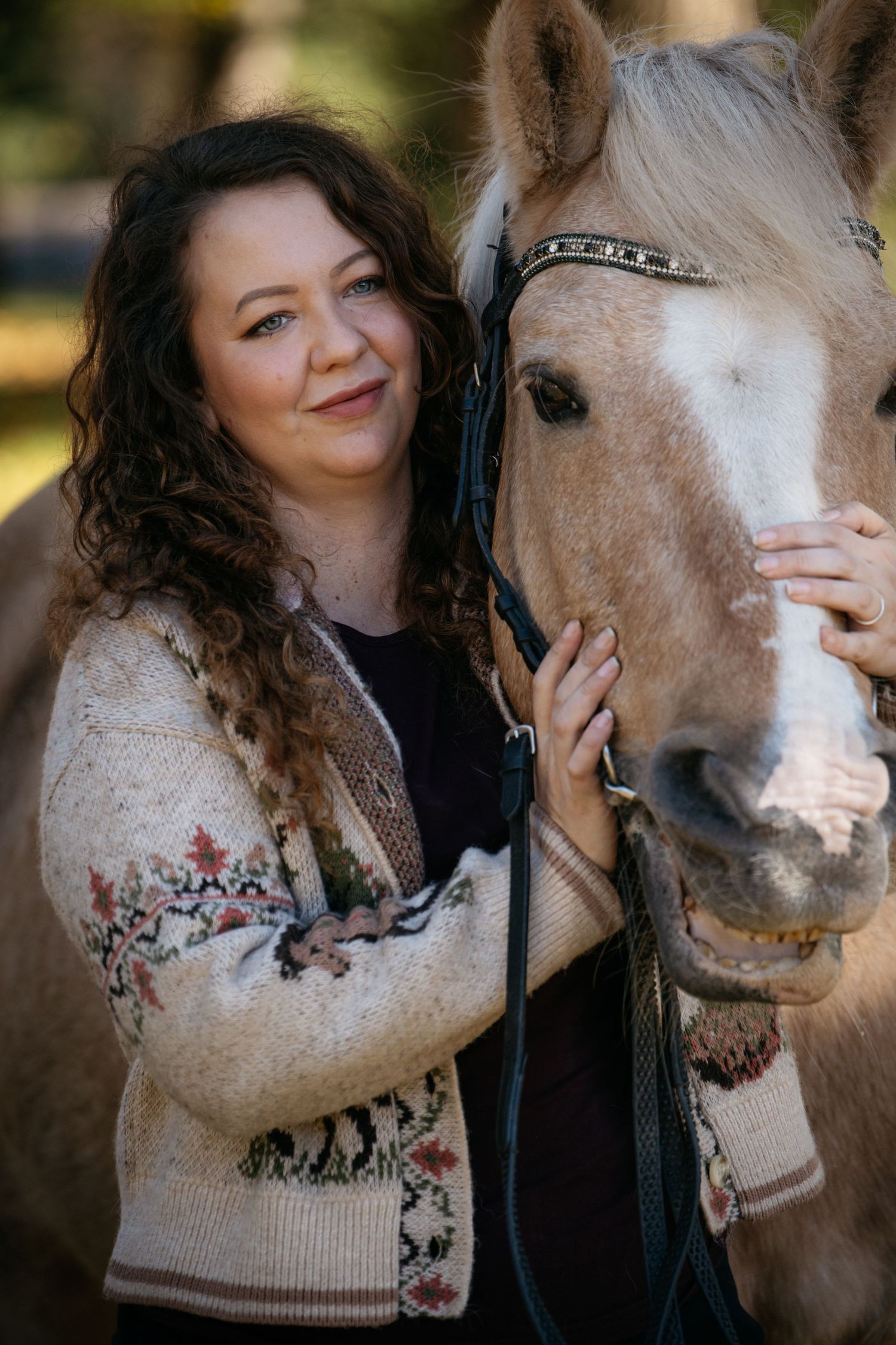 Woman with curly hair in floral cardigan smiling beside a palomino horse outdoors.