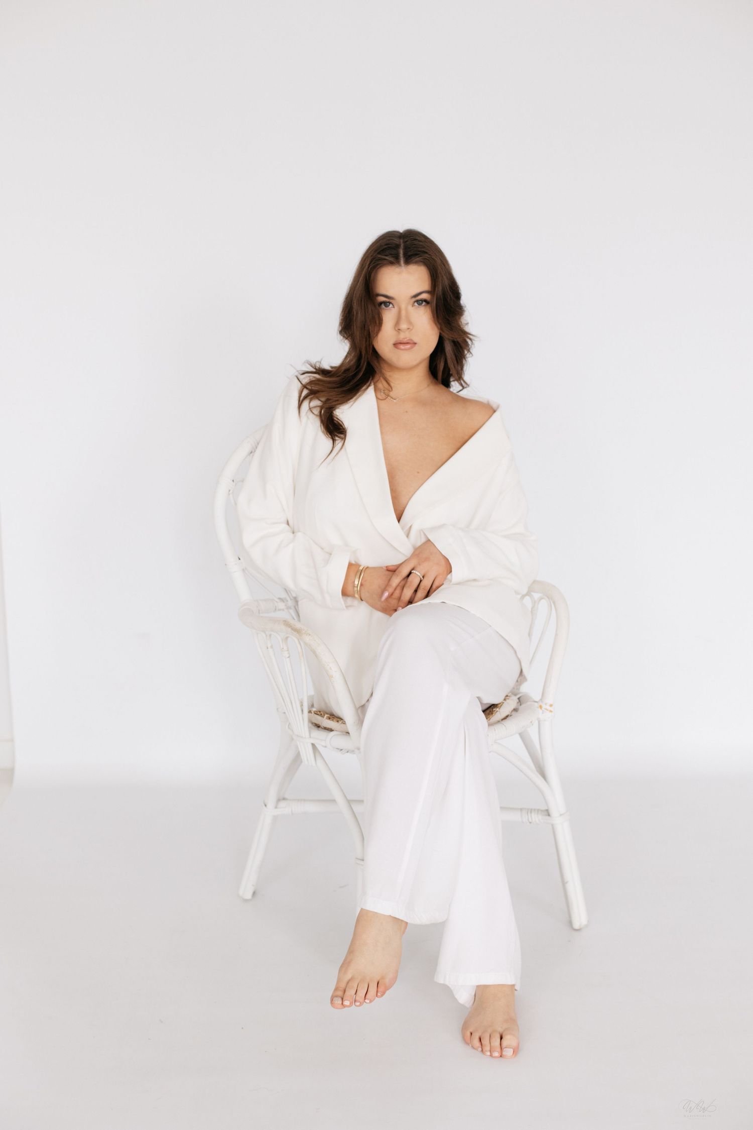 A brunette woman in an all-white outfit sits barefoot in a wicker chair against a white background.