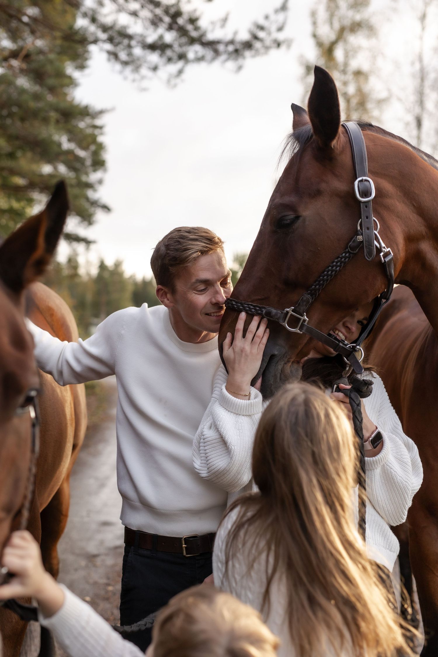 A man in a white sweater gently pets a brown horse outdoors near a forest with a woman beside him.
