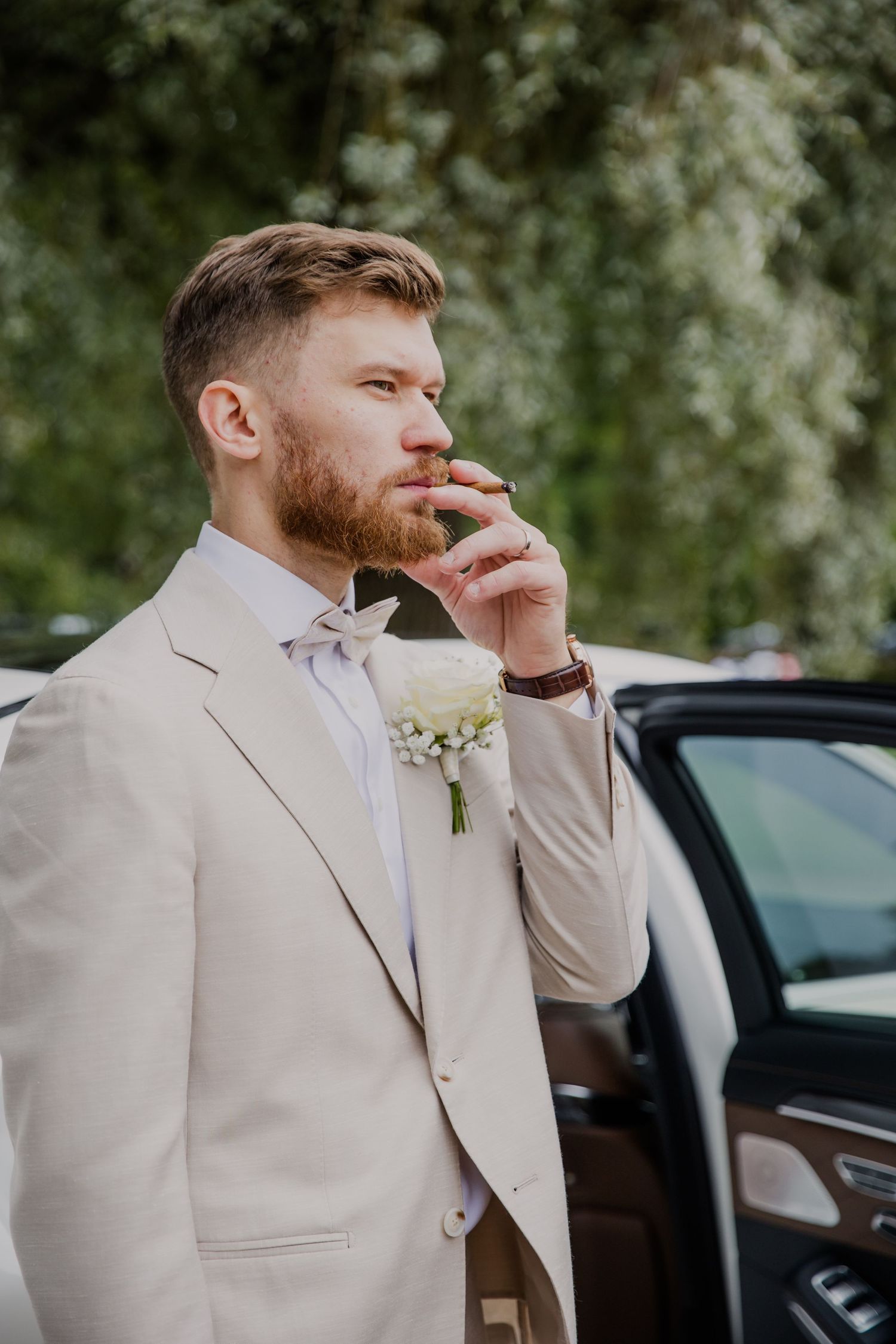Handsome groom in beige suit with white boutonniere standing by a car, thoughtfully touching his beard.