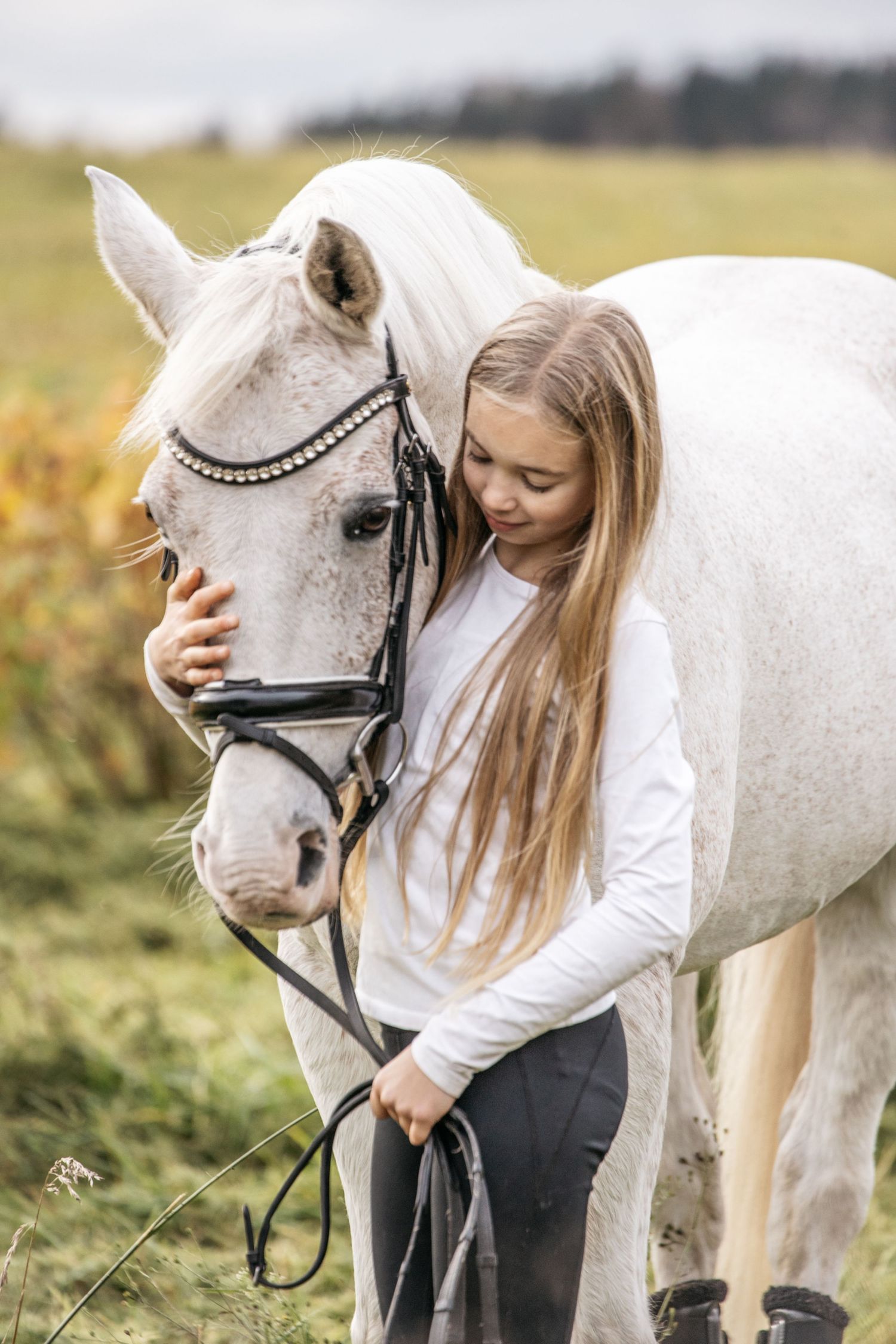 A young blonde girl in a white shirt tenderly embraces a white horse with a bridle in a green field.
