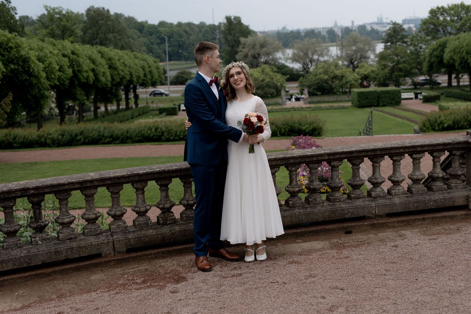 Wedding couple embracing on stone terrace overlooking park, bride in white dress holding bouquet, groom in blue suit.