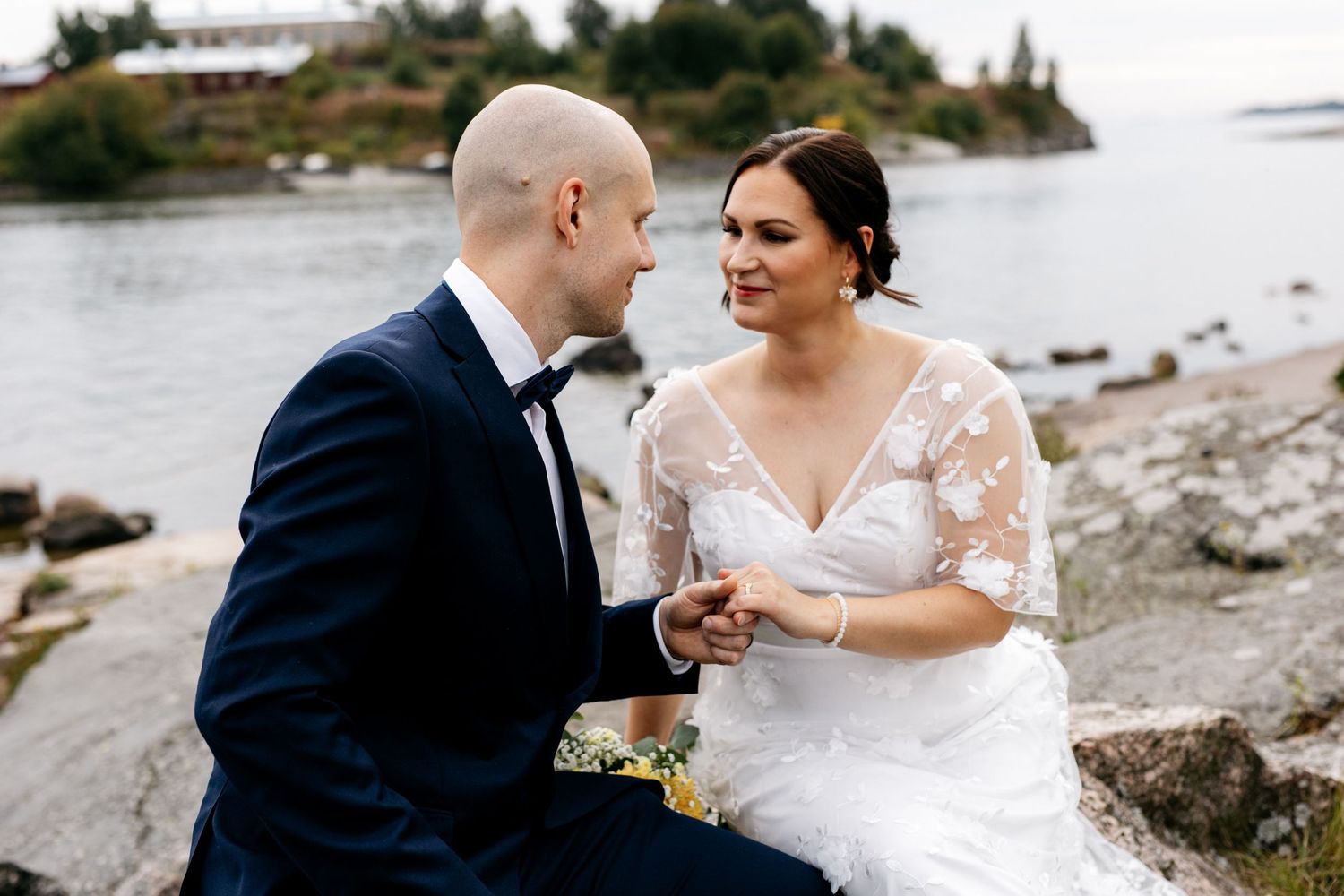 Bride and groom sitting on rocky waterside, holding hands and gazing at each other lovingly.