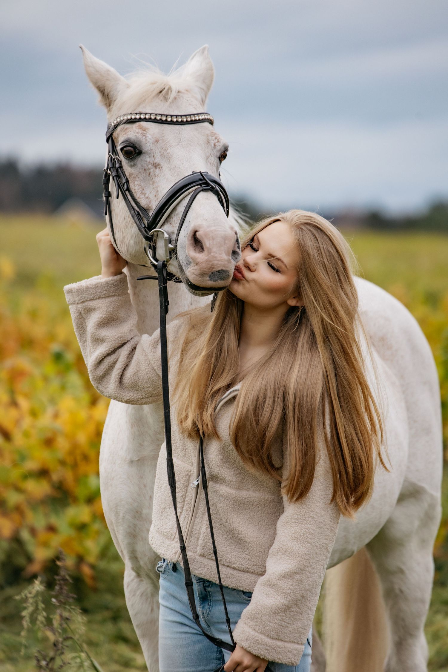 Young woman kissing a white horse in a field, wearing a beige sweater and jeans on an autumn day.