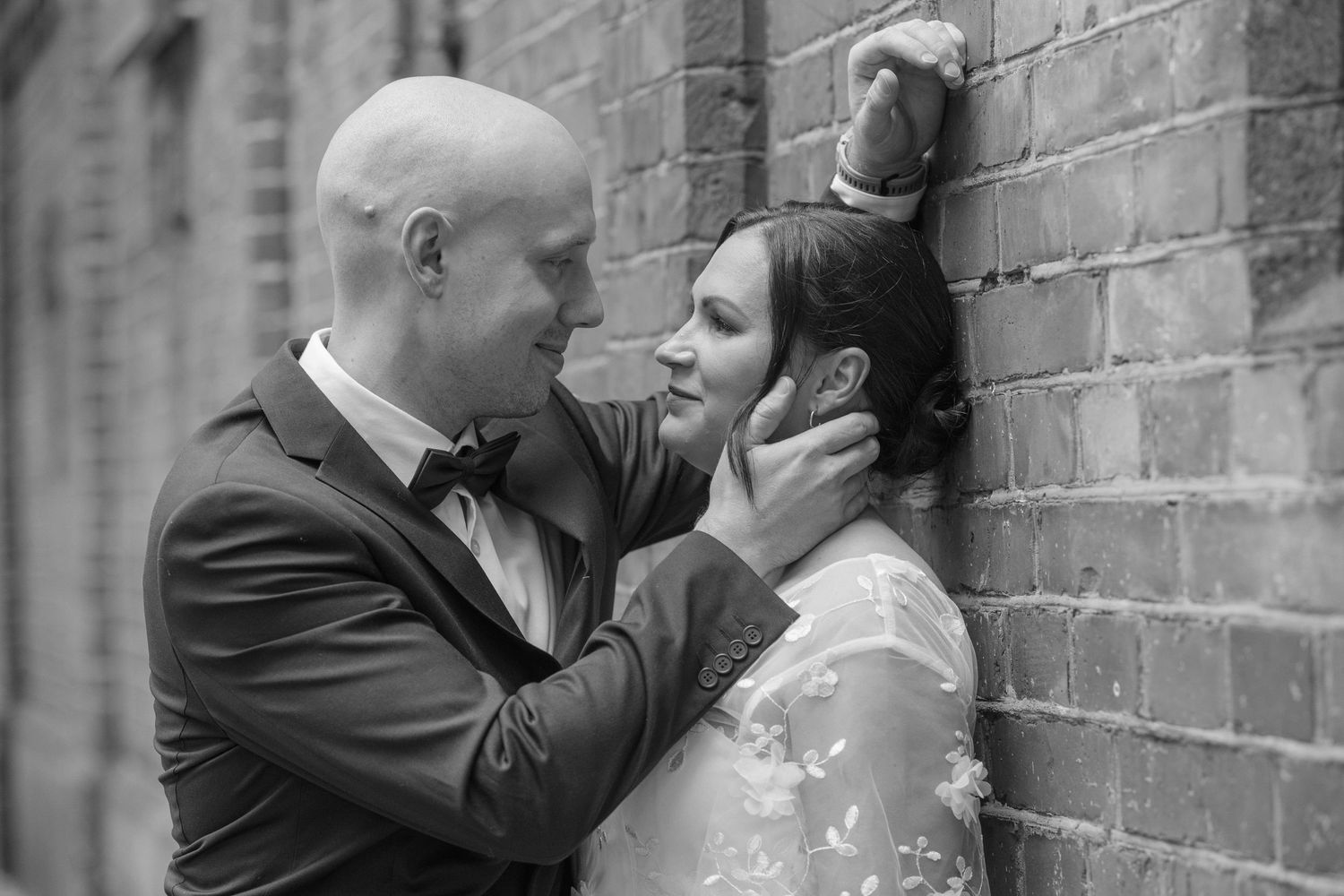 Black and white photo of a groom tenderly holding his bride's face against a brick wall.