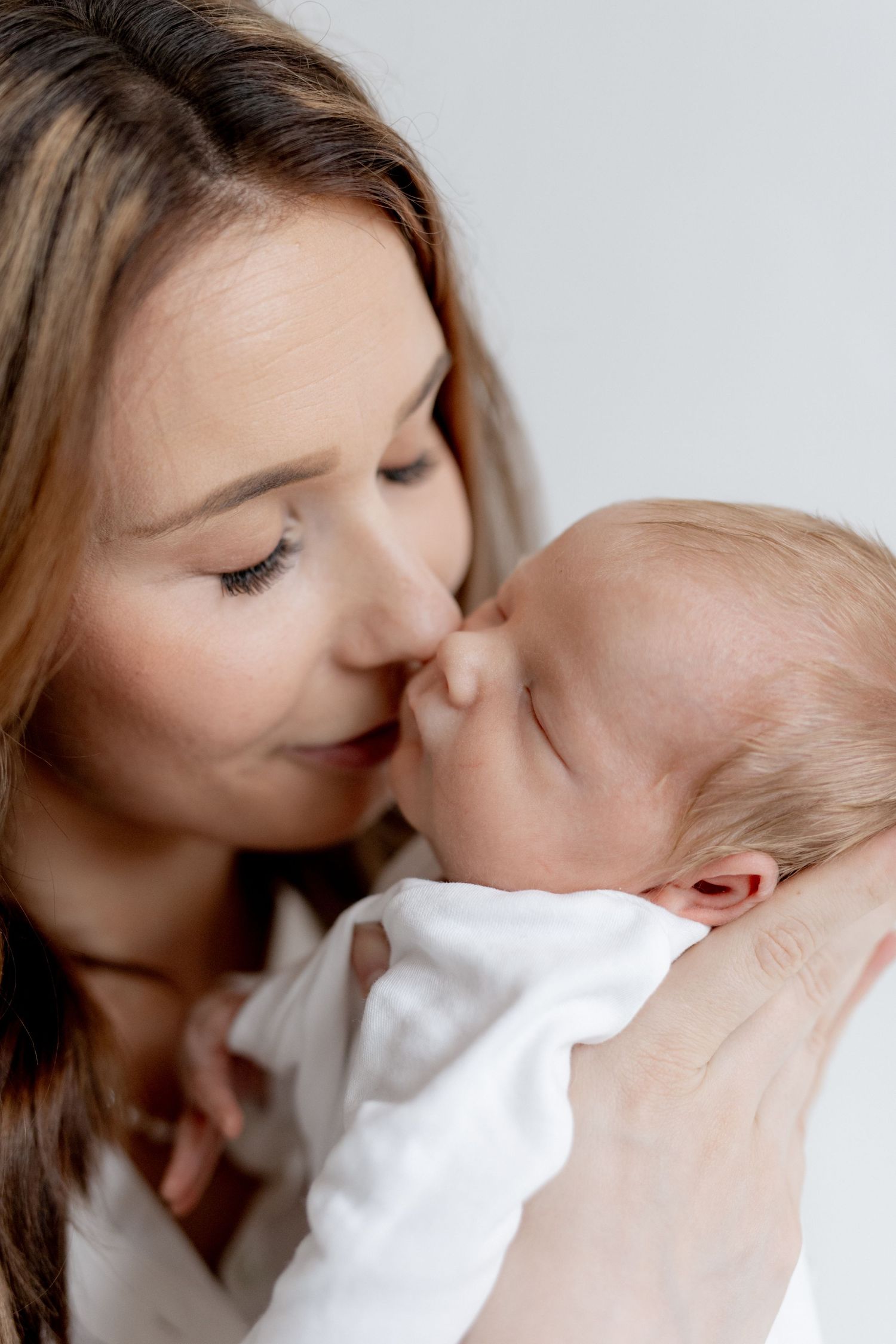 Smiling mother with long brown hair gently kisses the nose of her sleeping newborn baby wrapped in a soft white blanket.