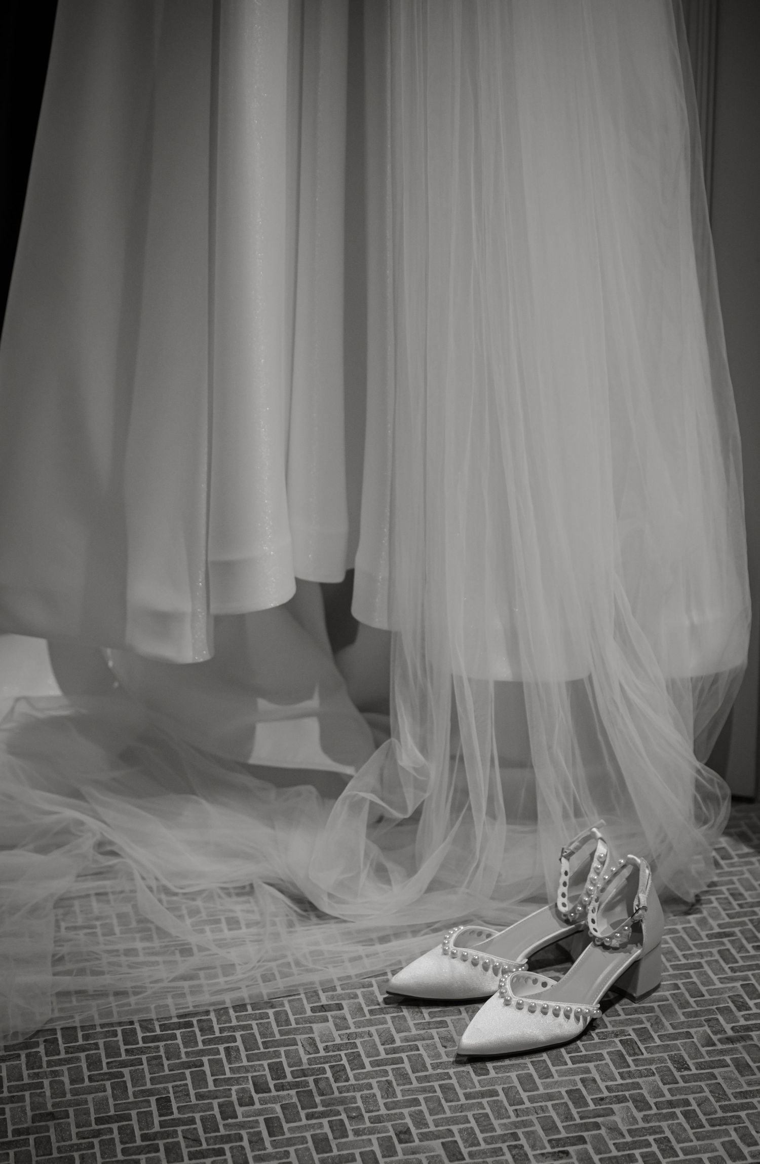 Black and white photo of white pearl-studded bridal heels placed beneath flowing tulle wedding gown.