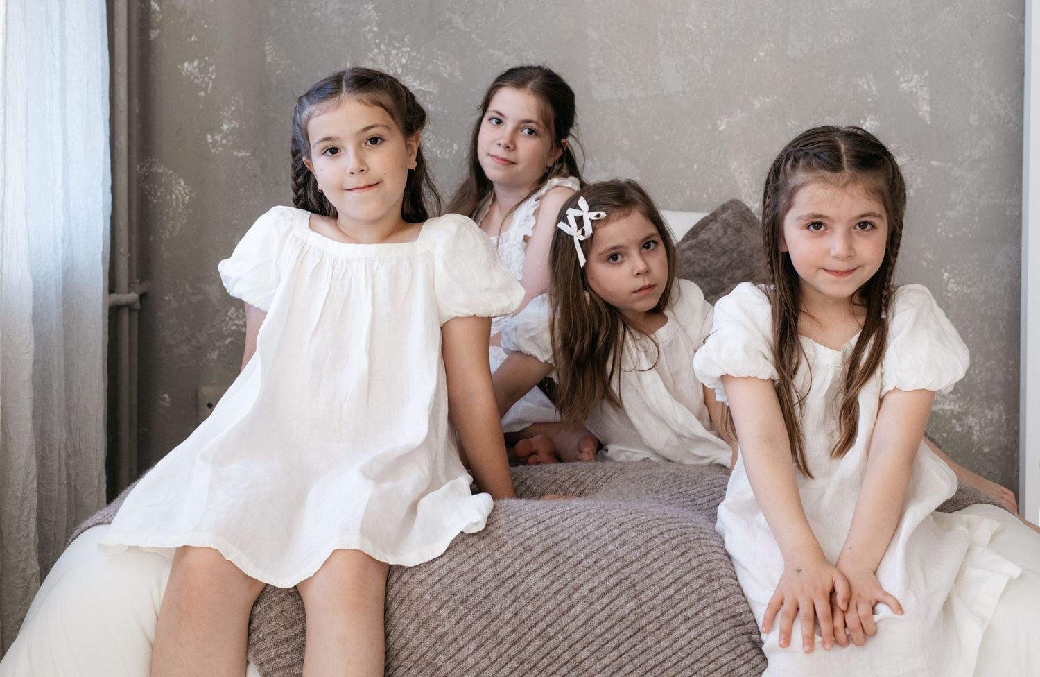 Four girls in white dresses sitting together on a bed in an elegant room.