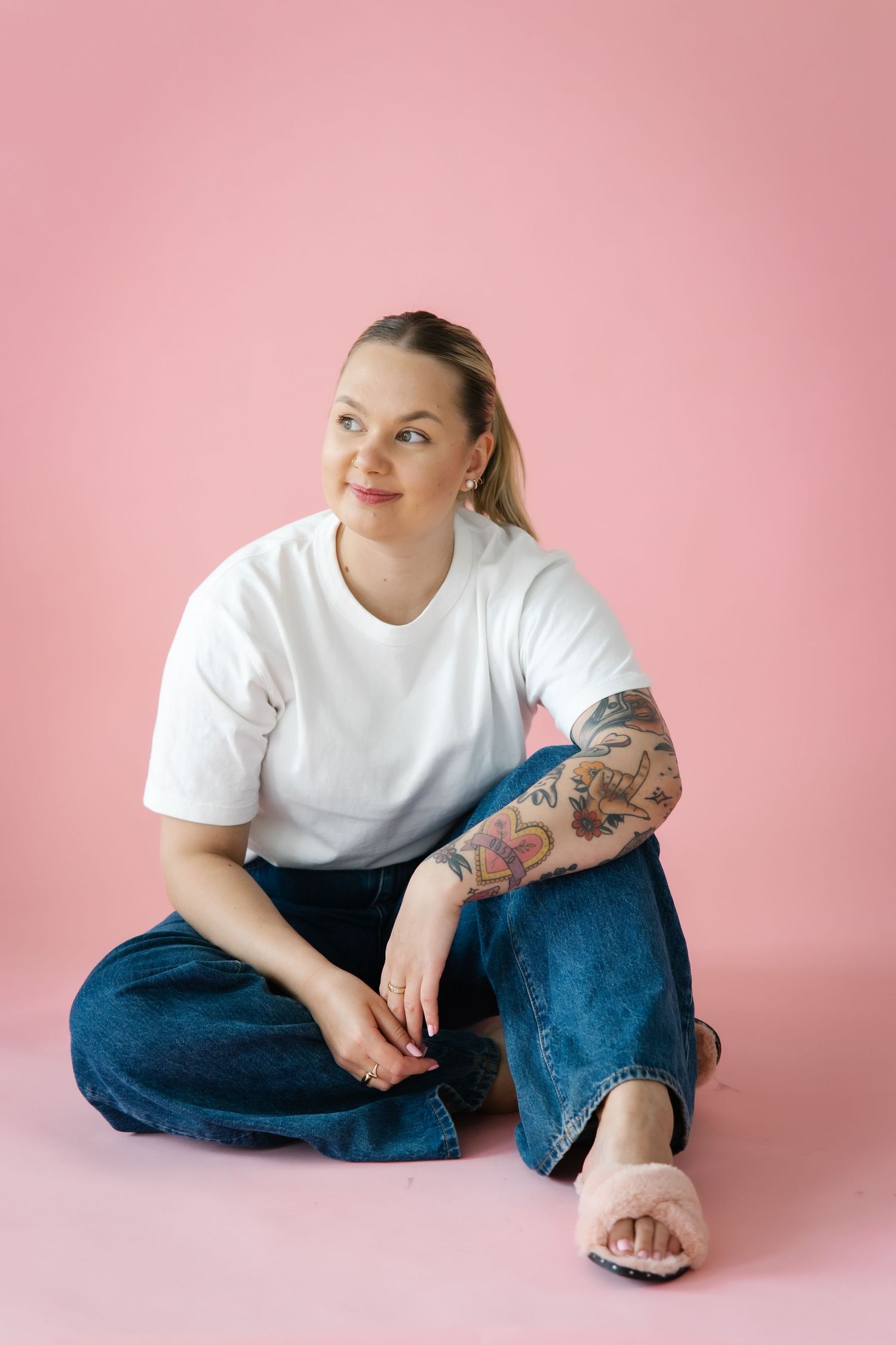 Woman with tattoo sleeve sits cross-legged in white tee and jeans on pink background.