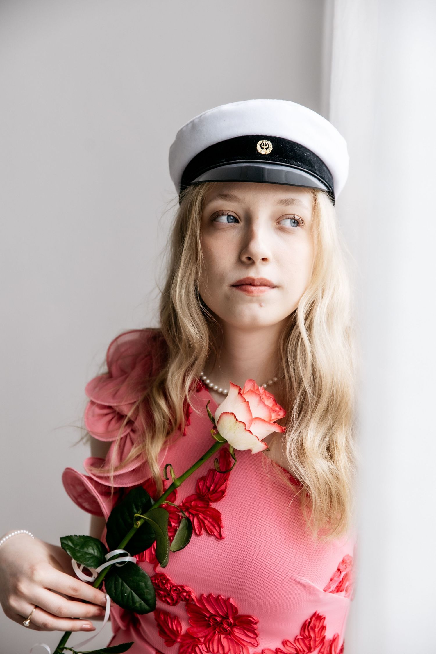 Young woman with long blonde hair in a white graduation cap and pink floral dress holds a pink rose while gazing out a window.