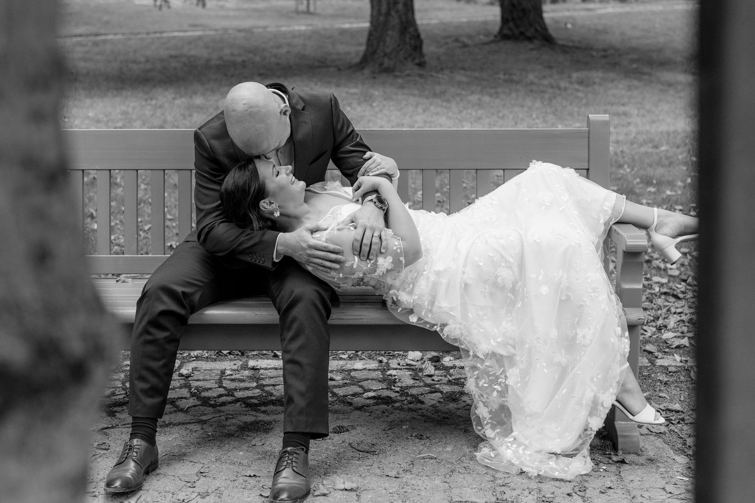 Black and white photo of a bride and groom sharing a romantic kiss on a park bench in their wedding attire.