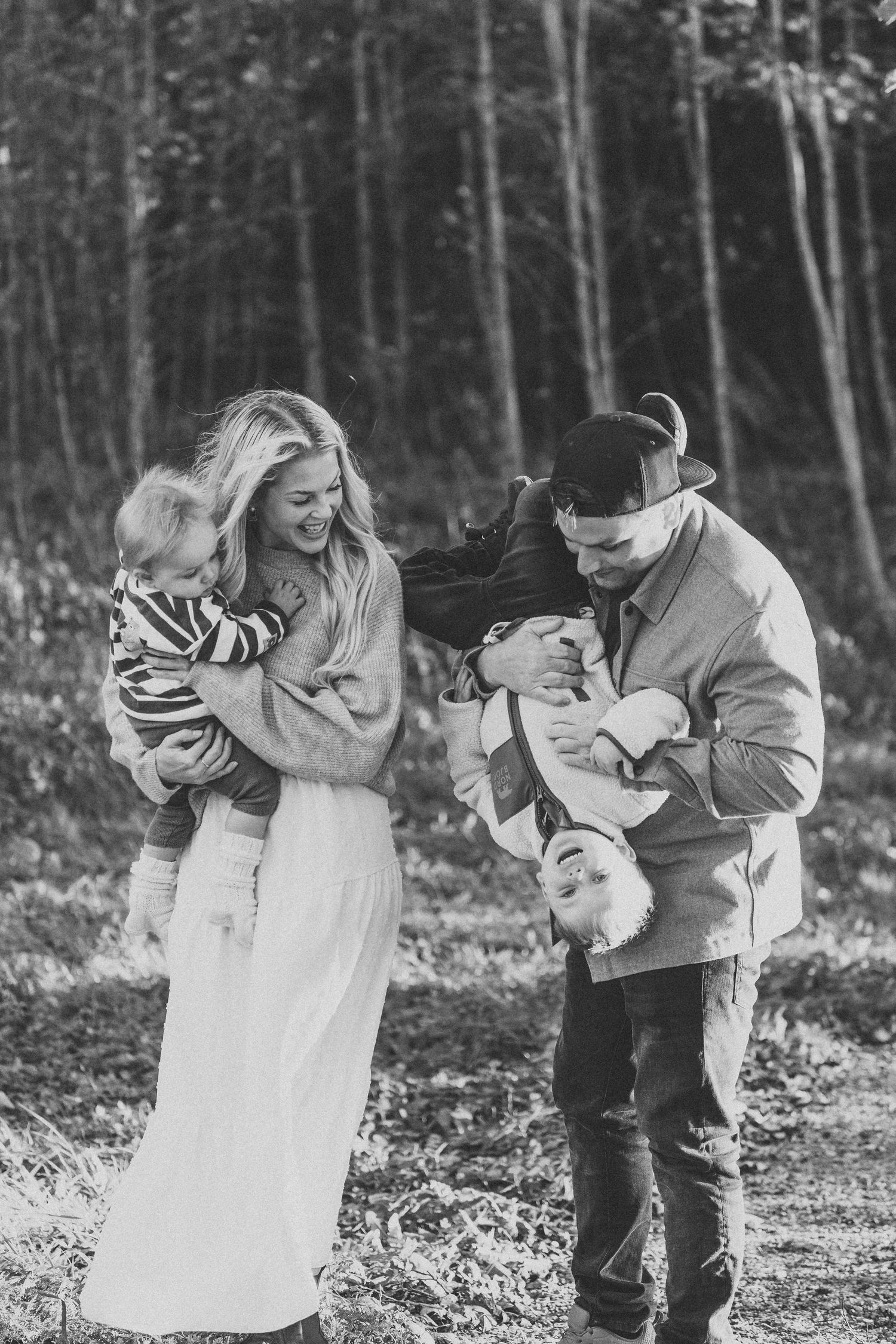 Black and white photo of a smiling family playing in a forest, with a mother holding a toddler and a father flipping a child upside down.