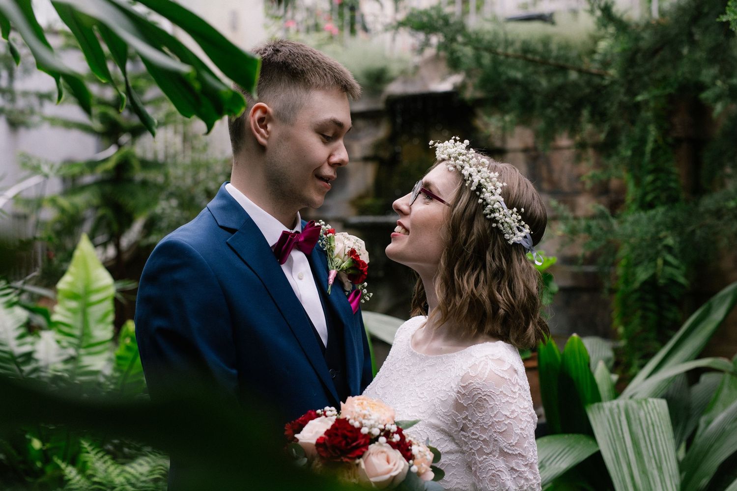 Wedding couple gazing at each other surrounded by tropical plants, bride in lace dress with floral crown.