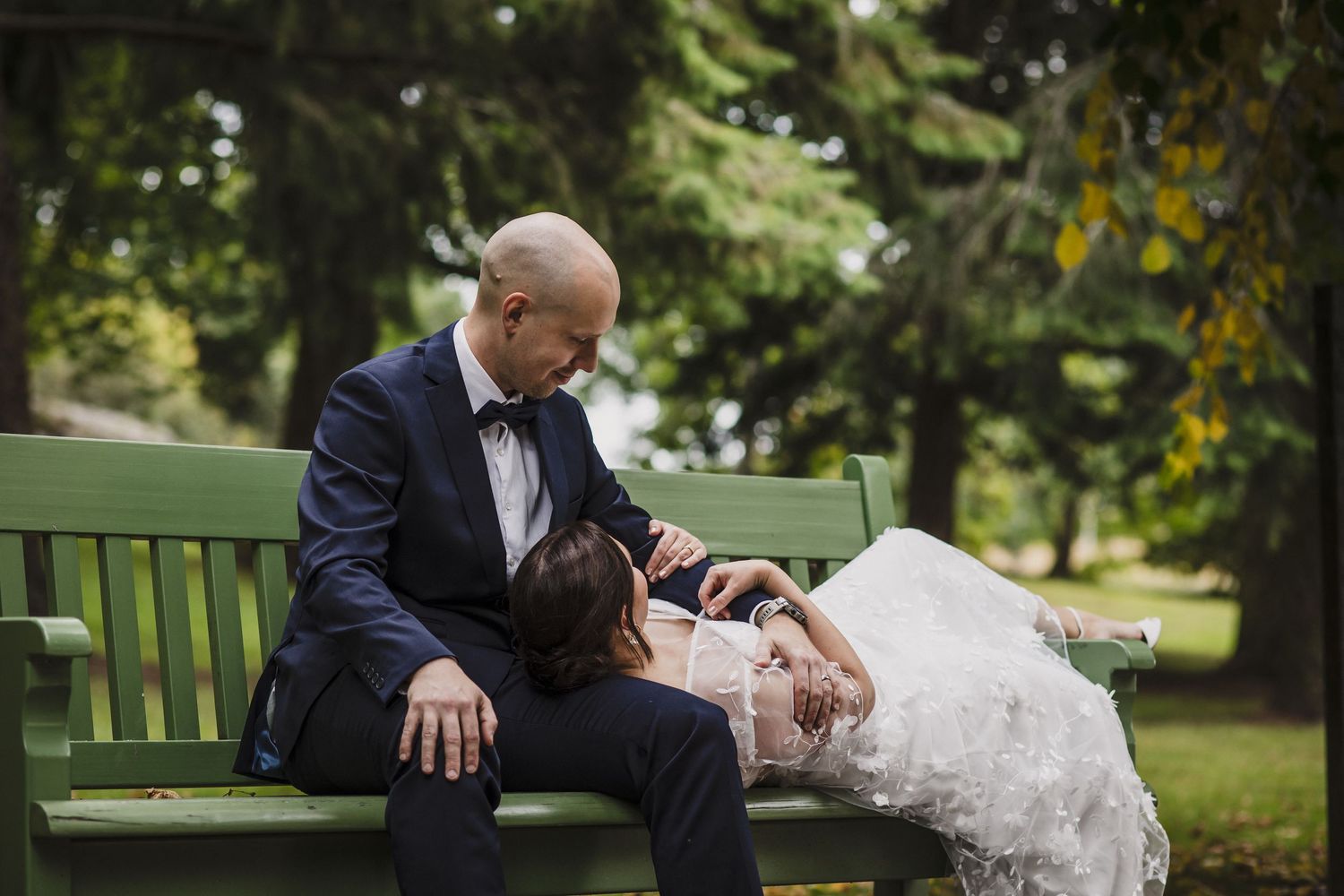 Groom in navy suit sitting on green park bench leaning toward bride lying across his lap.