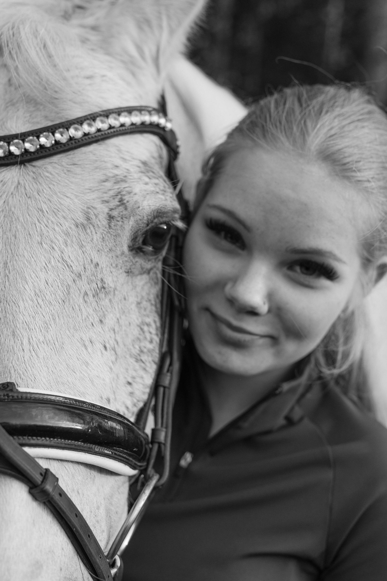 Black and white portrait of a young blonde woman cheek to cheek with a white horse wearing a rhinestone bridle.
