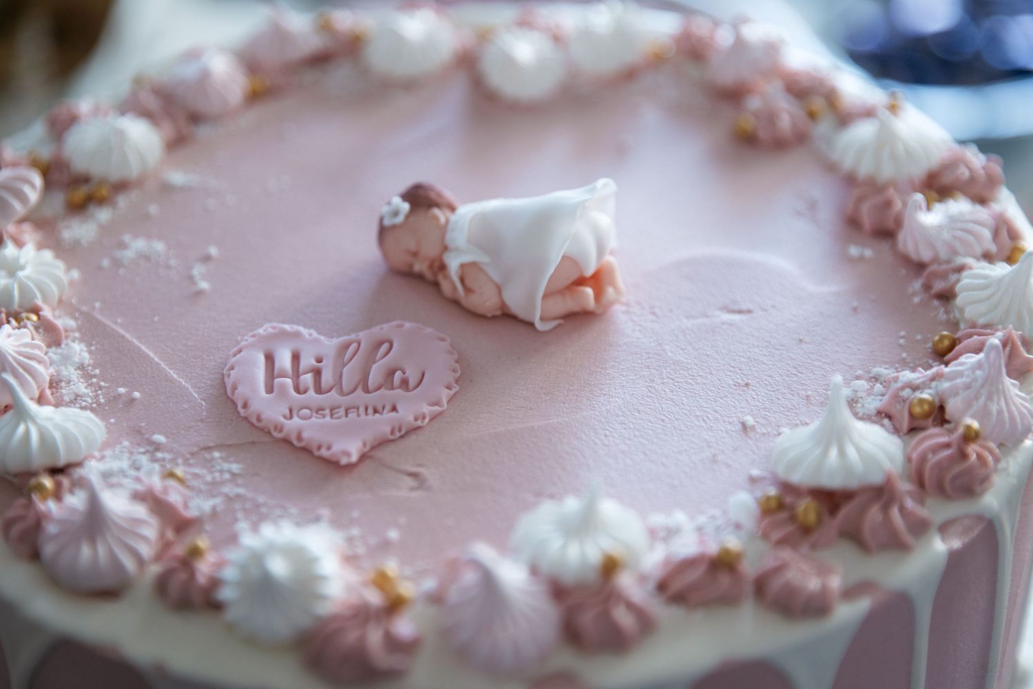 Pink christening cake with baby figurine and heart plaque reading Hilla, decorated with meringues and pink frosting.
