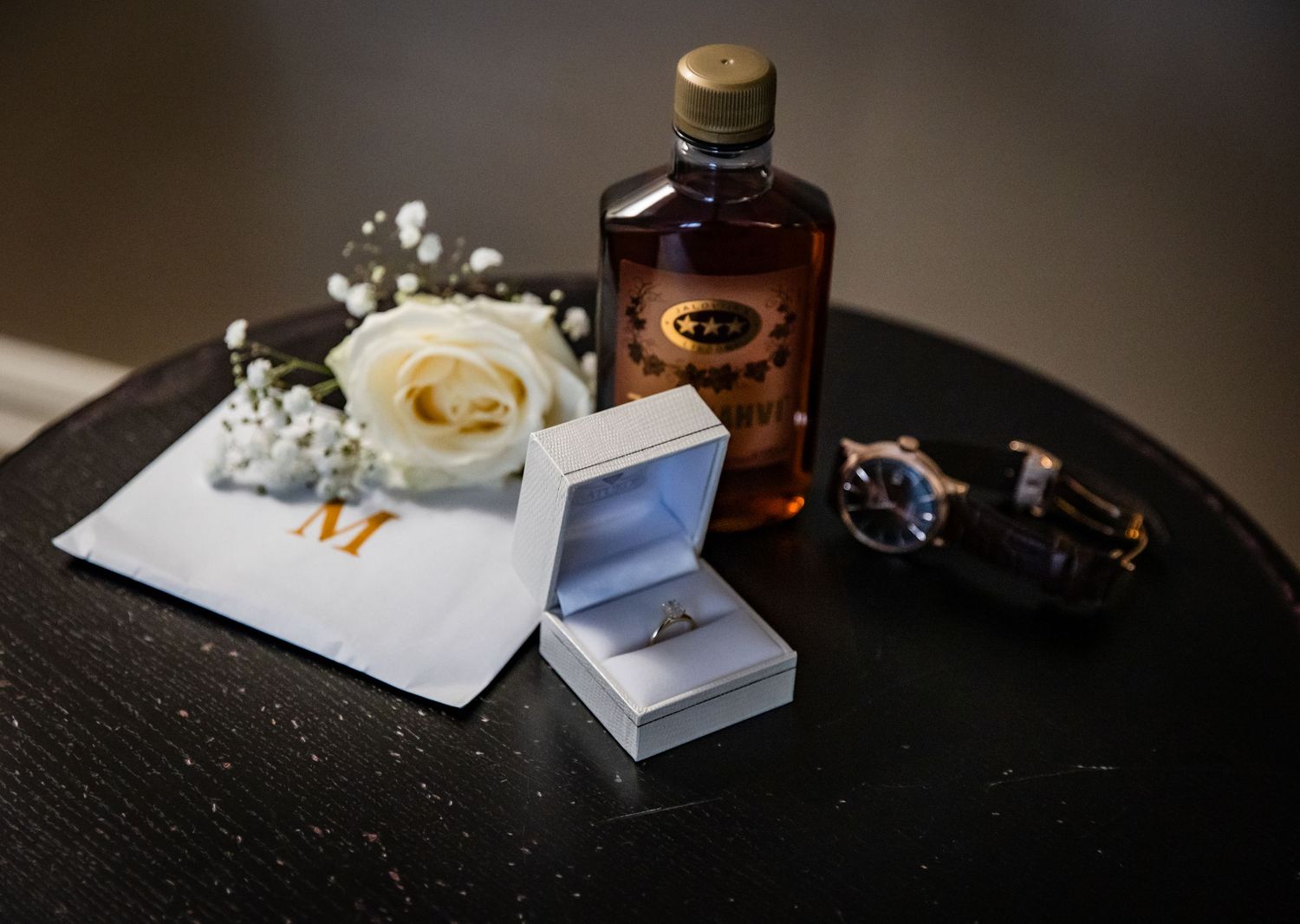 Wedding flat lay with ring box, white rose boutonniere, whiskey bottle, watch, and monogrammed handkerchief on dark table.