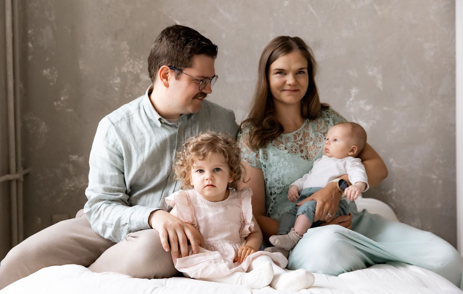 Happy family of four with toddler and newborn baby sitting together on bed against gray wall.
