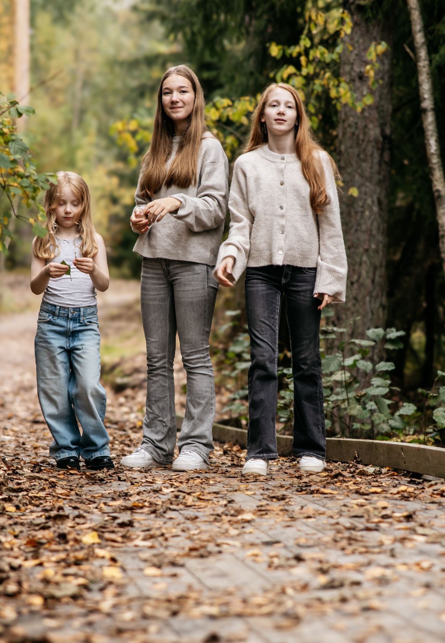 Three girls stand on a leaf-covered path in an autumn forest, wearing casual gray and beige outfits.