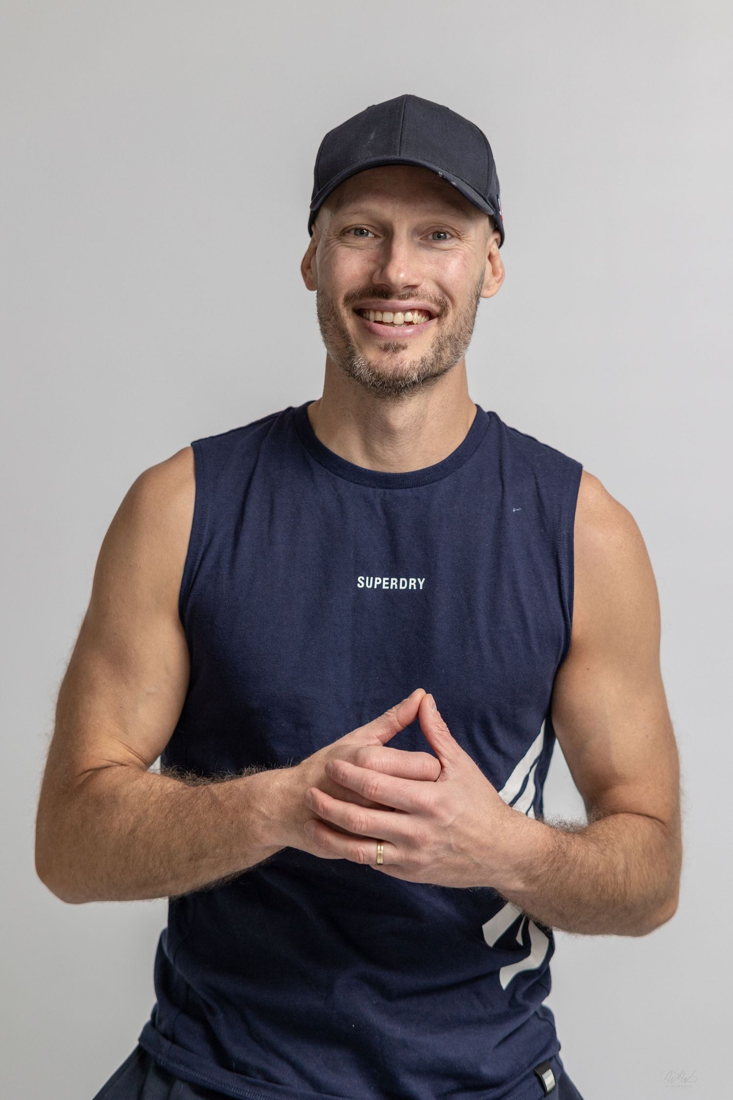 Smiling man in navy Superdry sleeveless shirt and cap with hands clasped, posing against gray background.