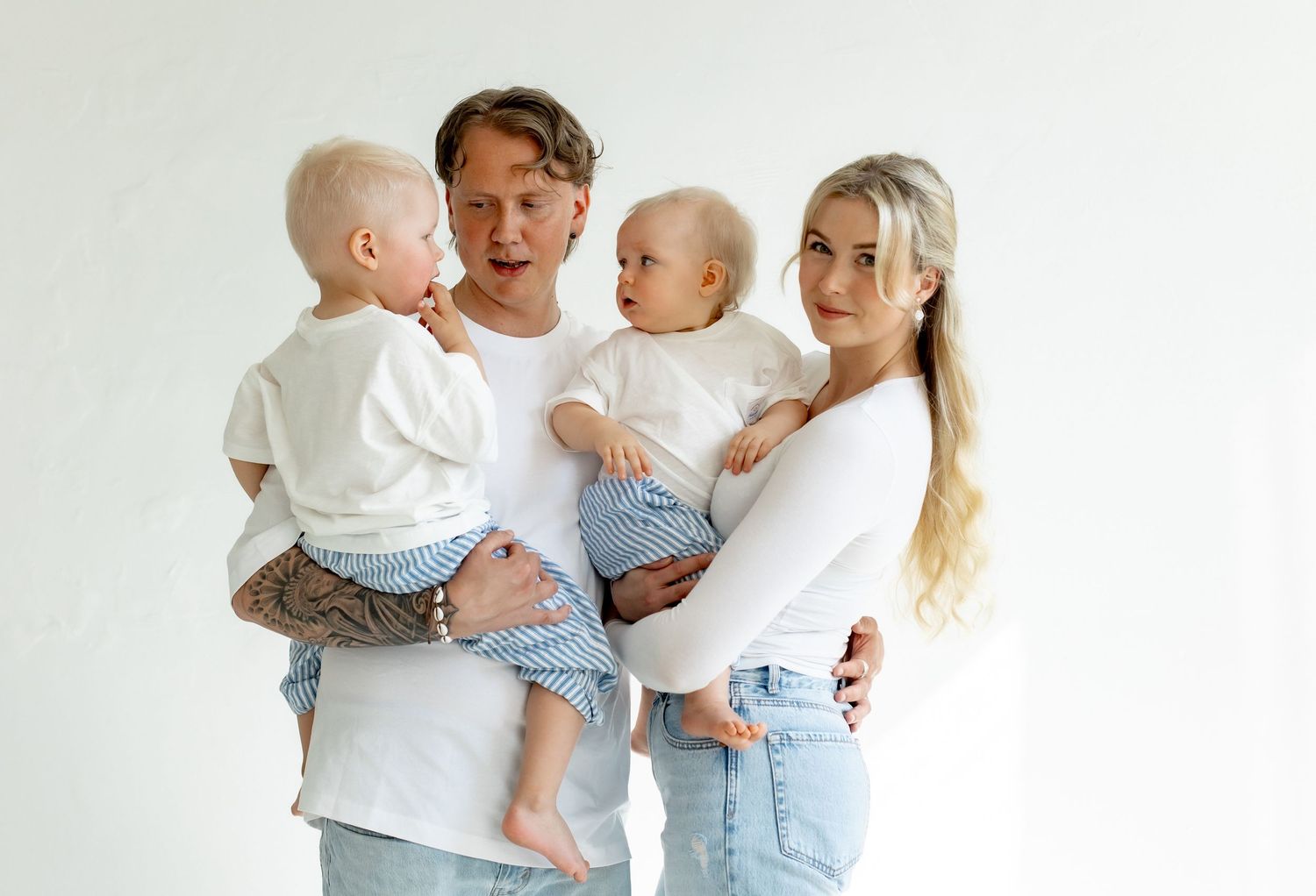 Young couple in white and denim outfits hold two toddlers against white background.