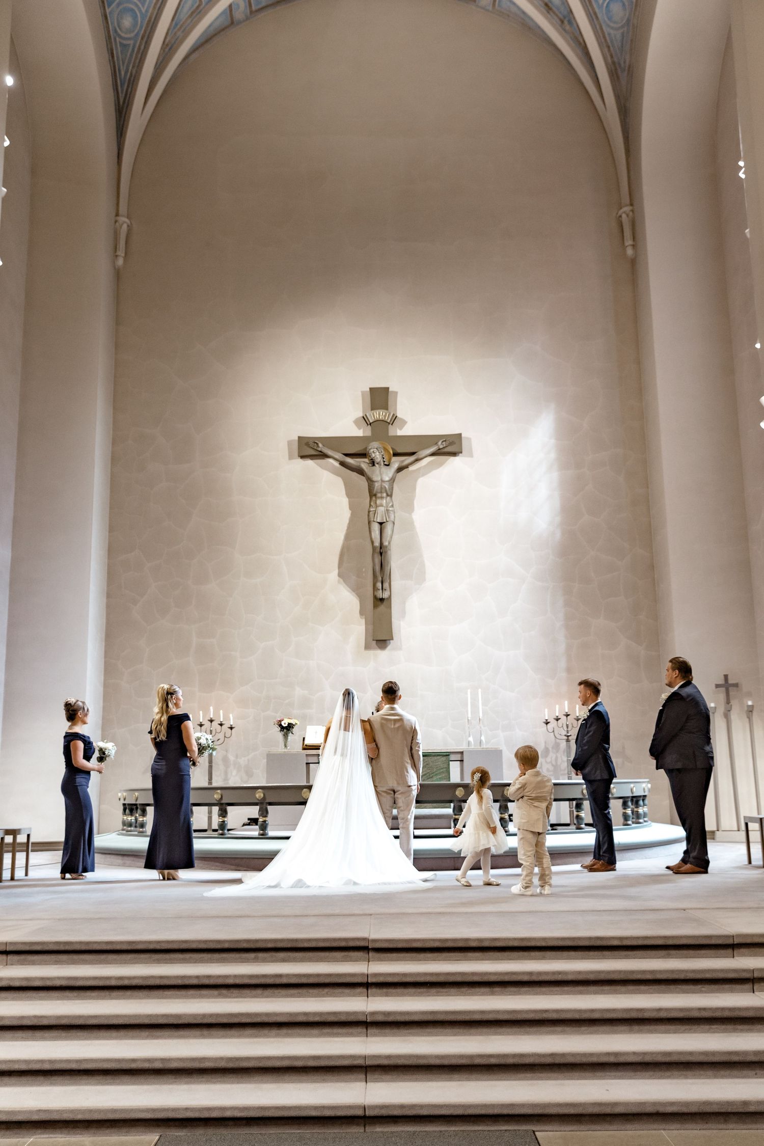 Bride and groom standing at church altar beneath crucifix during wedding ceremony with guests and flower girl present.