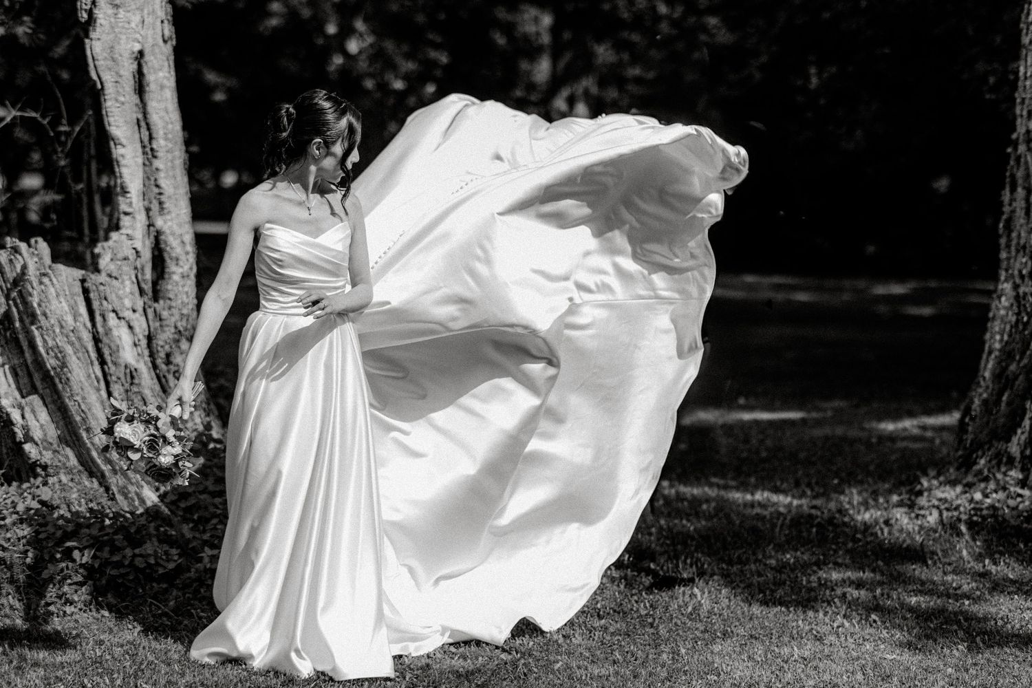 Black and white photo of bride in white gown with skirt billowing in wind among trees outdoors.